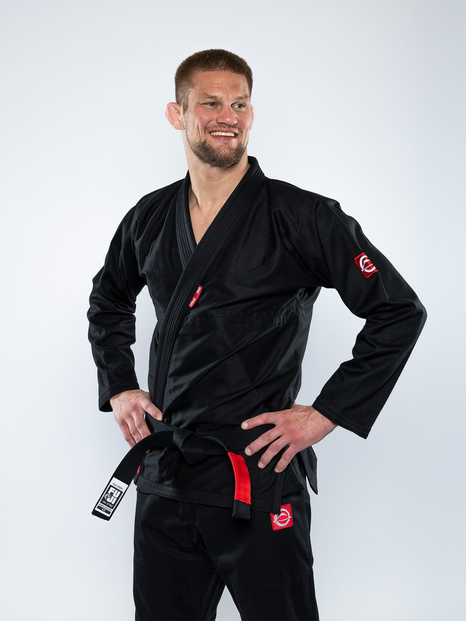 A man stands confidently with hands on hips, smiling in the Ultra Lite 300 BJJ Gi Black with red accents and a black belt, against a plain white background.