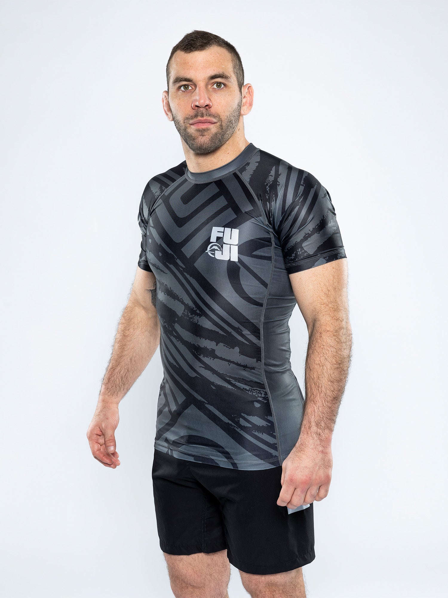 A man with short brown hair and a trimmed beard wears the Battle Flex Lite Rashguard Black, paired with black shorts, standing against a plain white background.