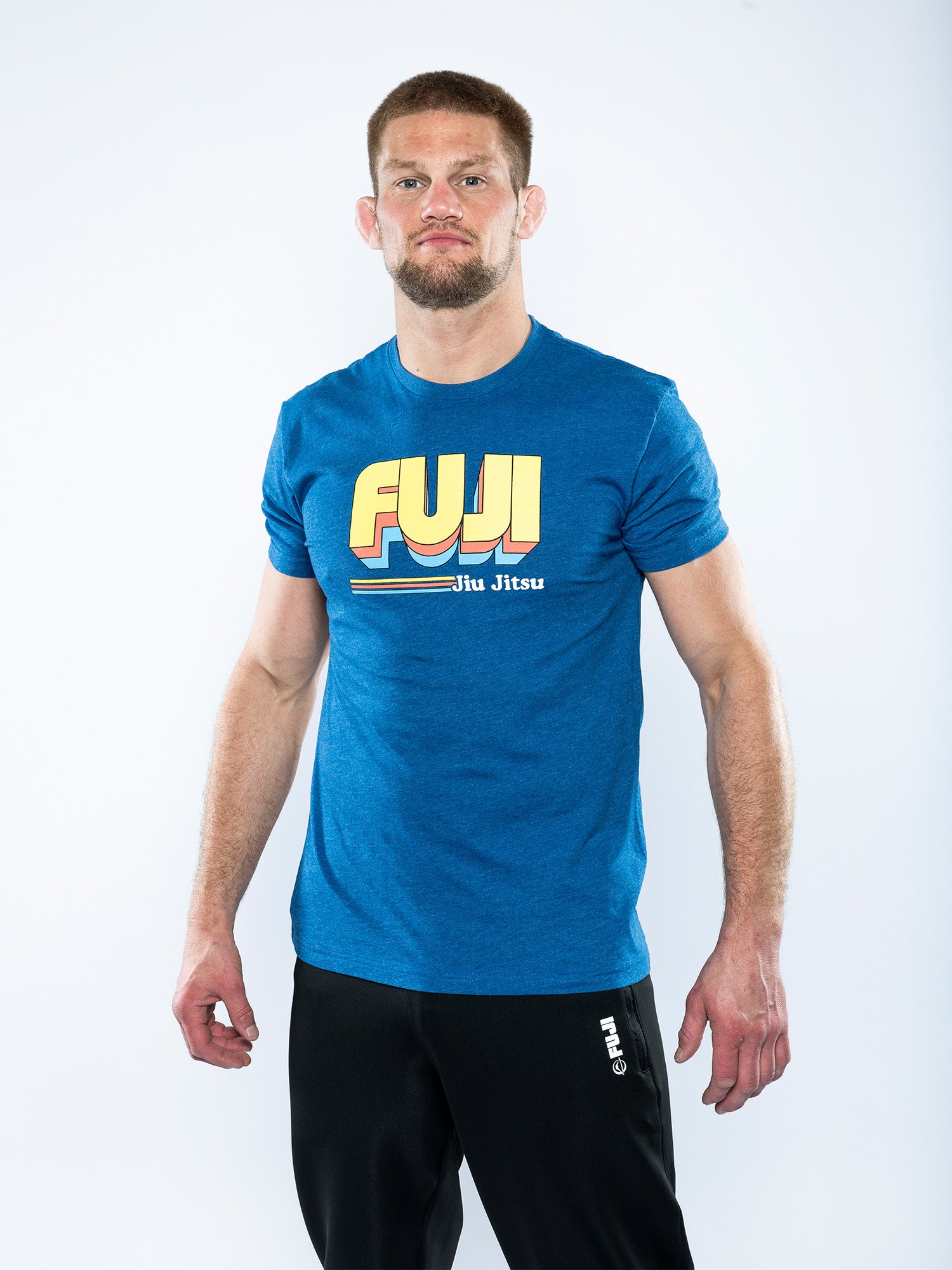 A man with short hair and a trimmed beard faces forward, wearing the Vintage Jiu Jitsu T-Shirt Blue featuring retro yellow lettering, paired with black athletic pants against a plain white background.