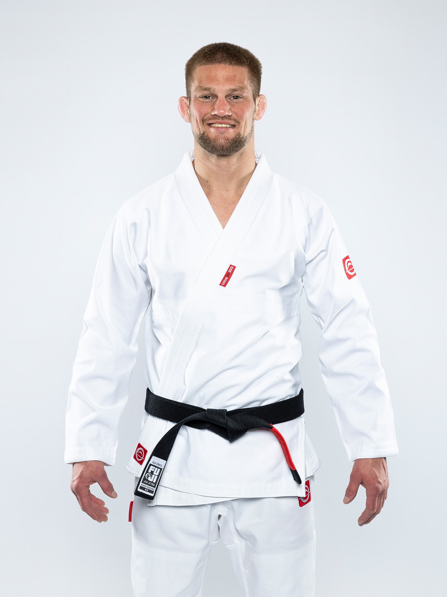 A man wearing the Ultra Lite 300 BJJ Gi White with a black belt stands facing forward and smiling against a plain, light background.