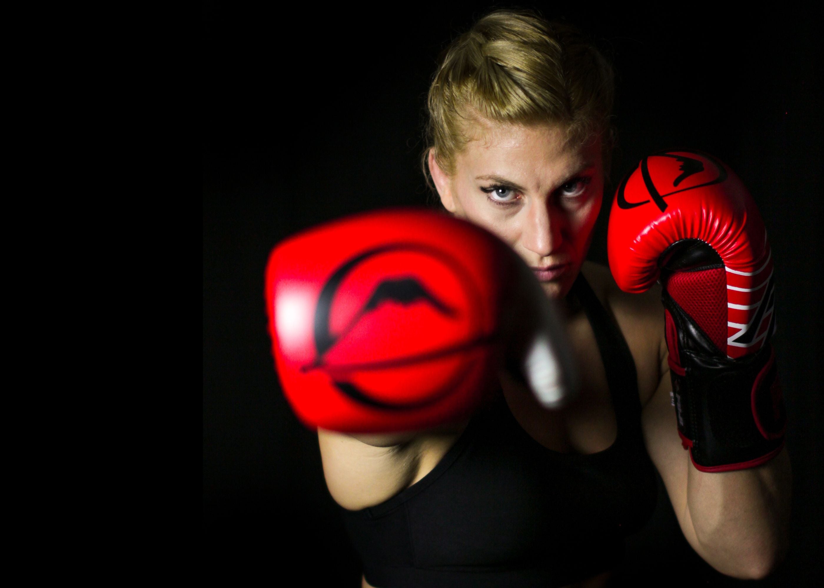 A woman with blonde hair in a braid, wearing red boxing gloves and a black sports bra, throws a punch towards the camera against a dark background.