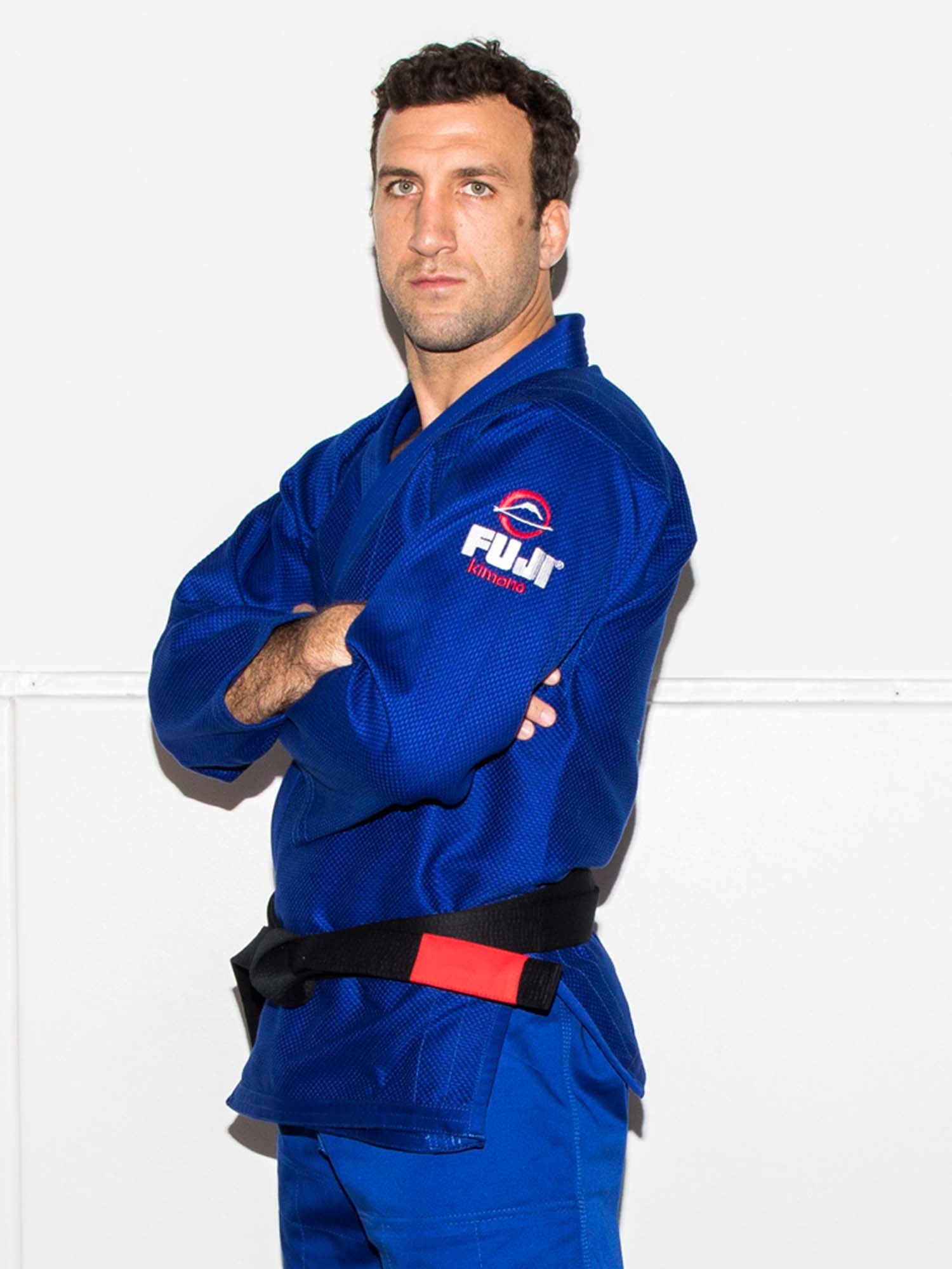 A man stands confidently with arms crossed against a white background, wearing the All Around BJJ Gi Blue with a FUJI logo and a black belt.