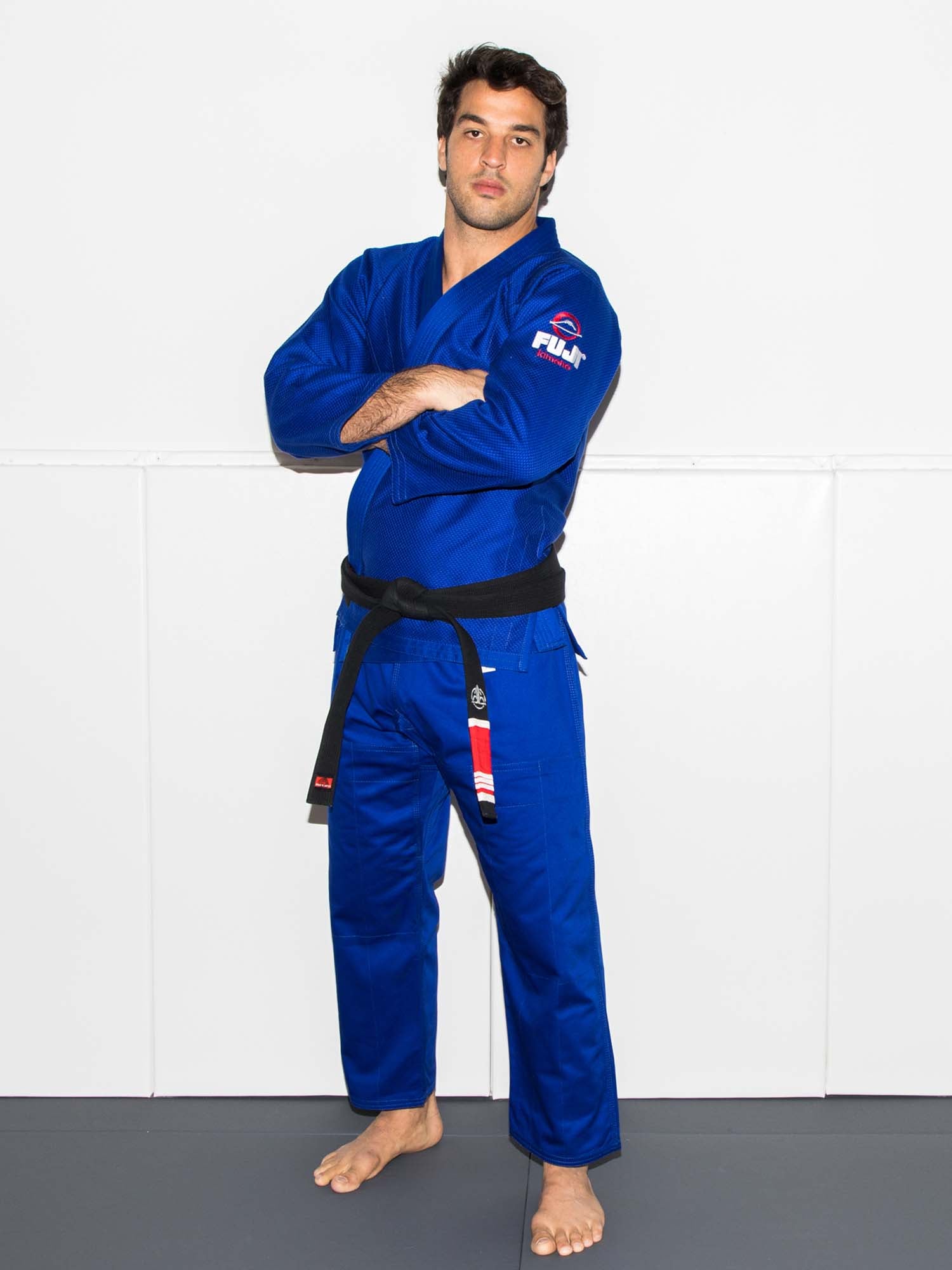A man wearing the All Around BJJ Gi Blue with a black belt stands barefoot on a mat, arms crossed, looking forward, with a white wall background.