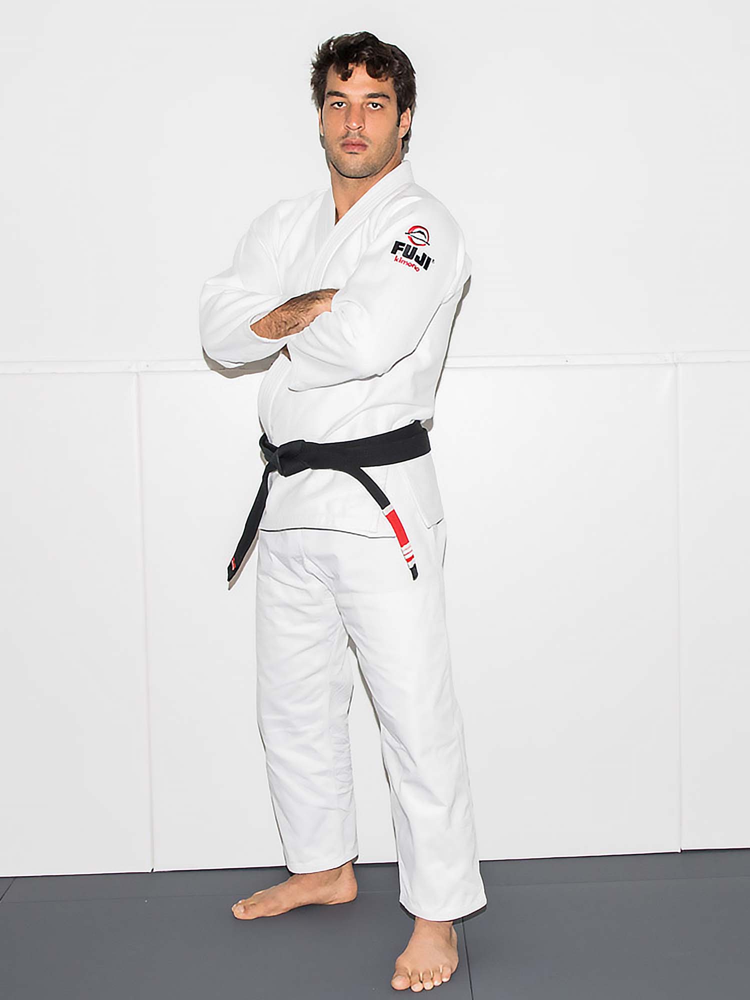 A man in the All Around BJJ Gi White with a black belt stands barefoot on a mat, arms crossed, facing the camera against a plain white background.
