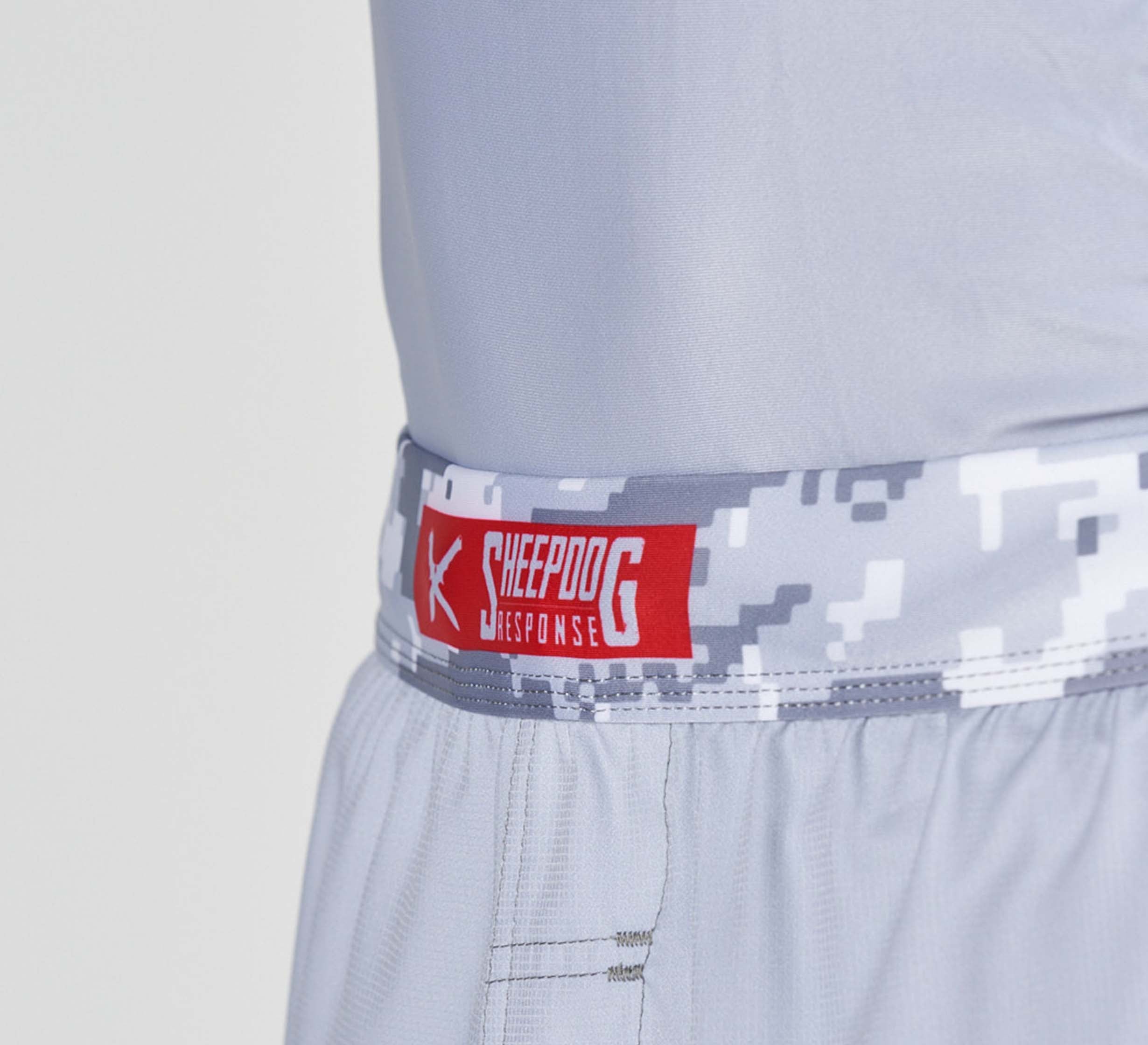 Close-up of a person wearing Sheepdog Response Flex Lite Shorts Grey, featuring a digital camouflage waistband with a red and white Sheepdog Response logo and text. The background is plain and light-colored.