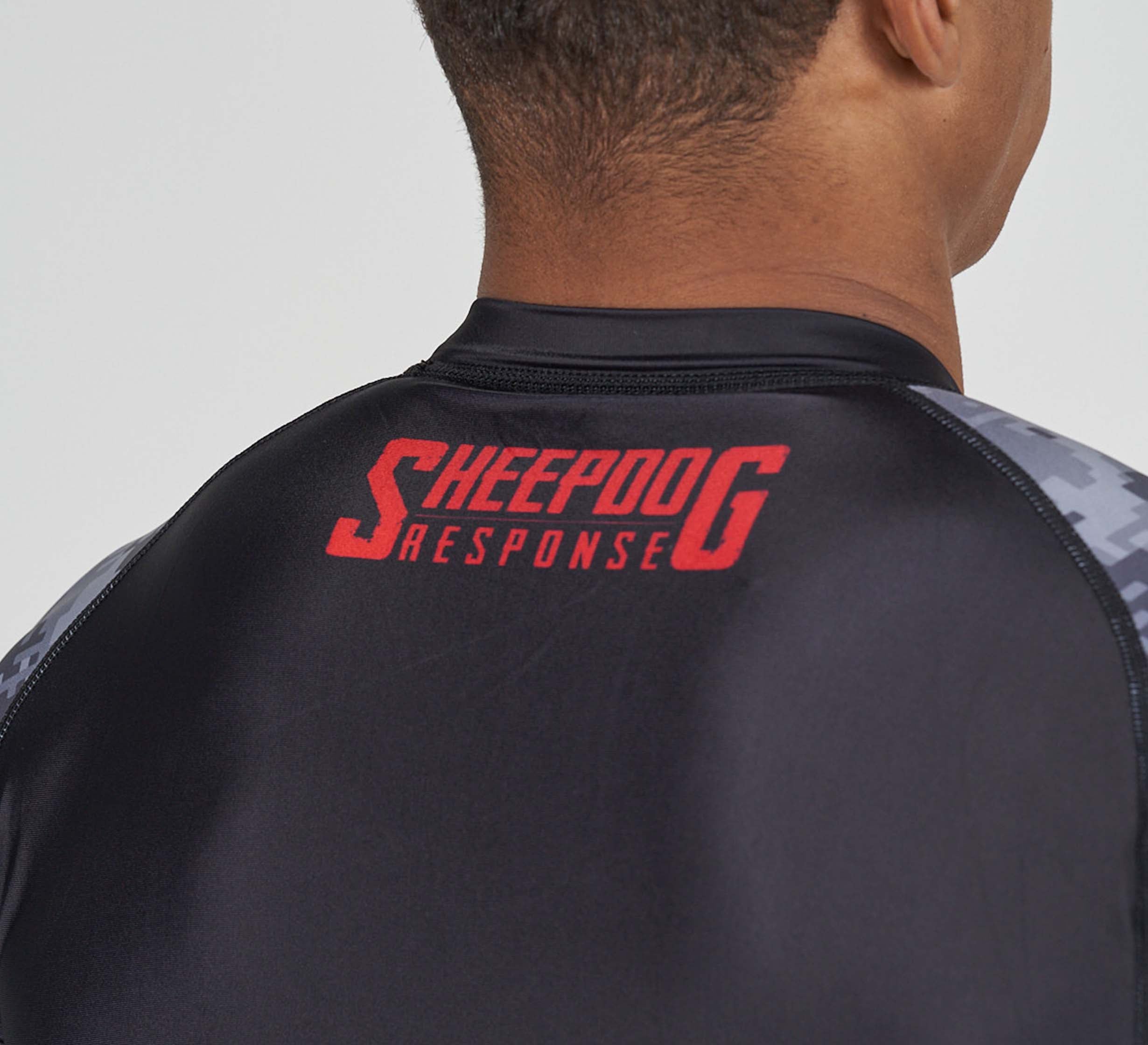 A person wears the Sheepdog Response Flex Lite Rashguard Black, featuring dark gray camo-patterned sleeves and large red "Sheepdog Response" lettering on the back of the shoulders.