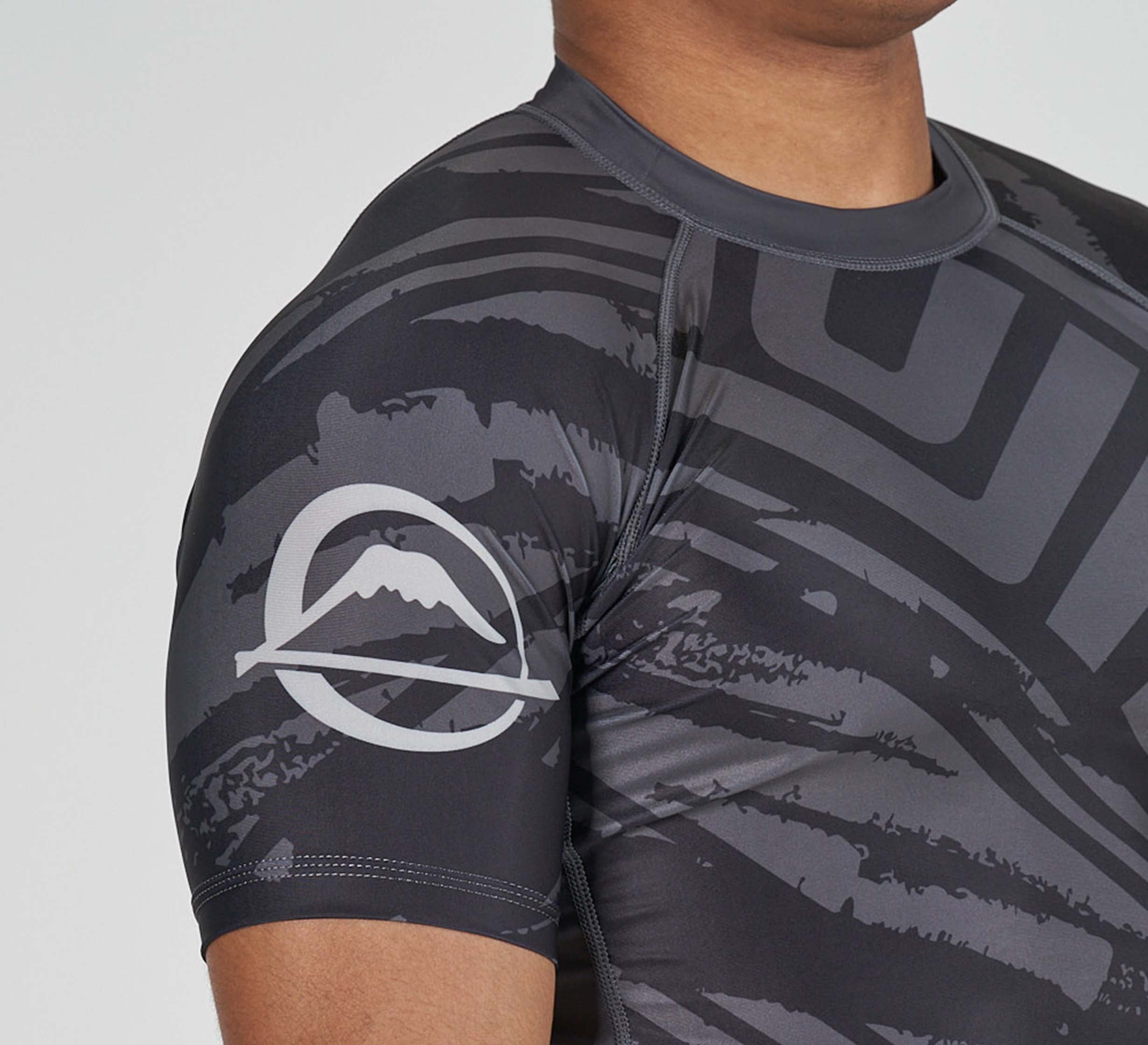 A person wears the Battle Flex Lite Rashguard Black, a short-sleeve athletic shirt with abstract black and gray patterns and a mountain logo on the sleeve, shown in a close-up of the shoulder and upper arm.