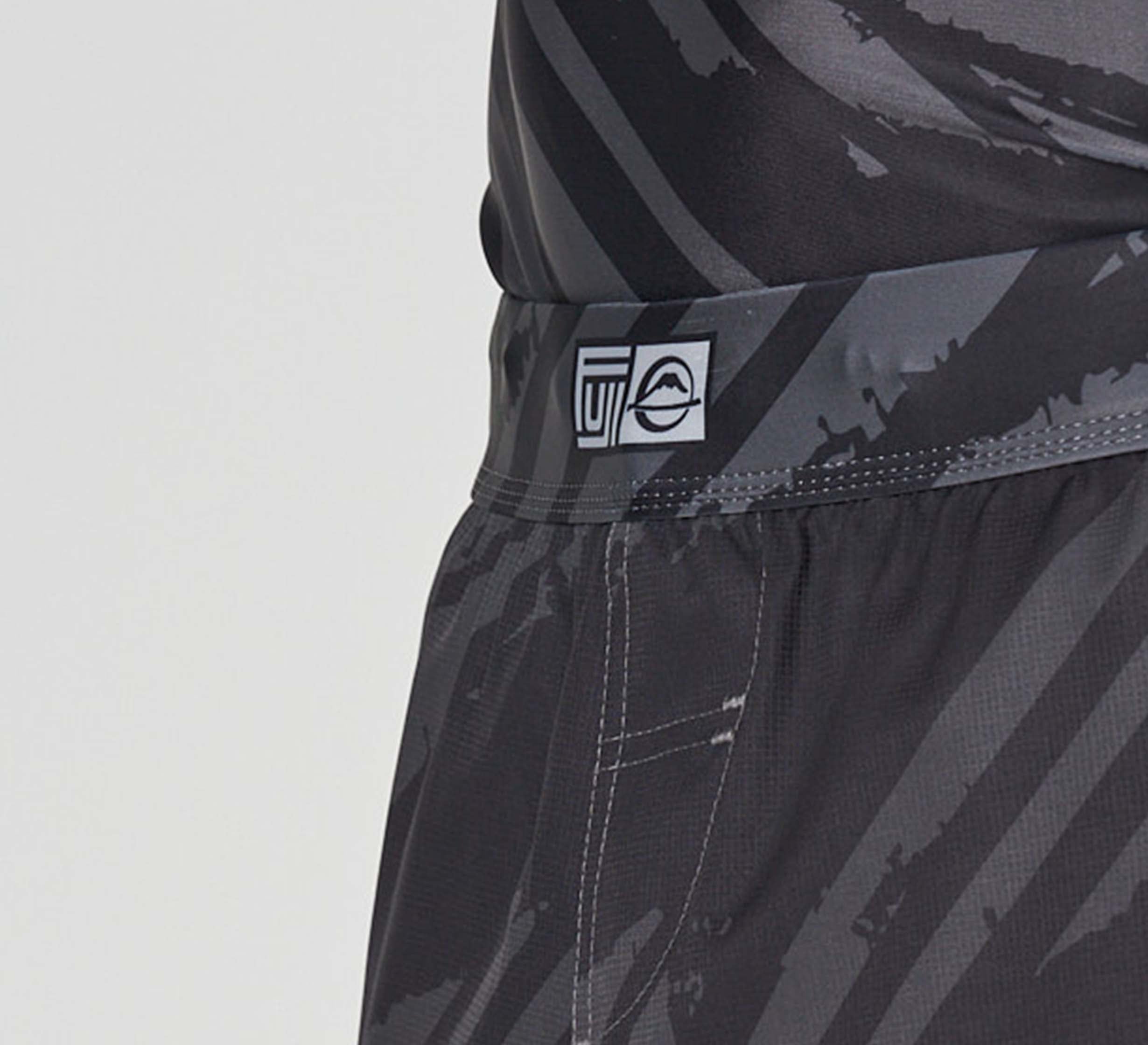 Close-up of a person wearing Battle Flex Lite Shorts Black—dark athletic shorts with black and gray diagonal patterns and a waistband with a small white tag featuring abstract logos.