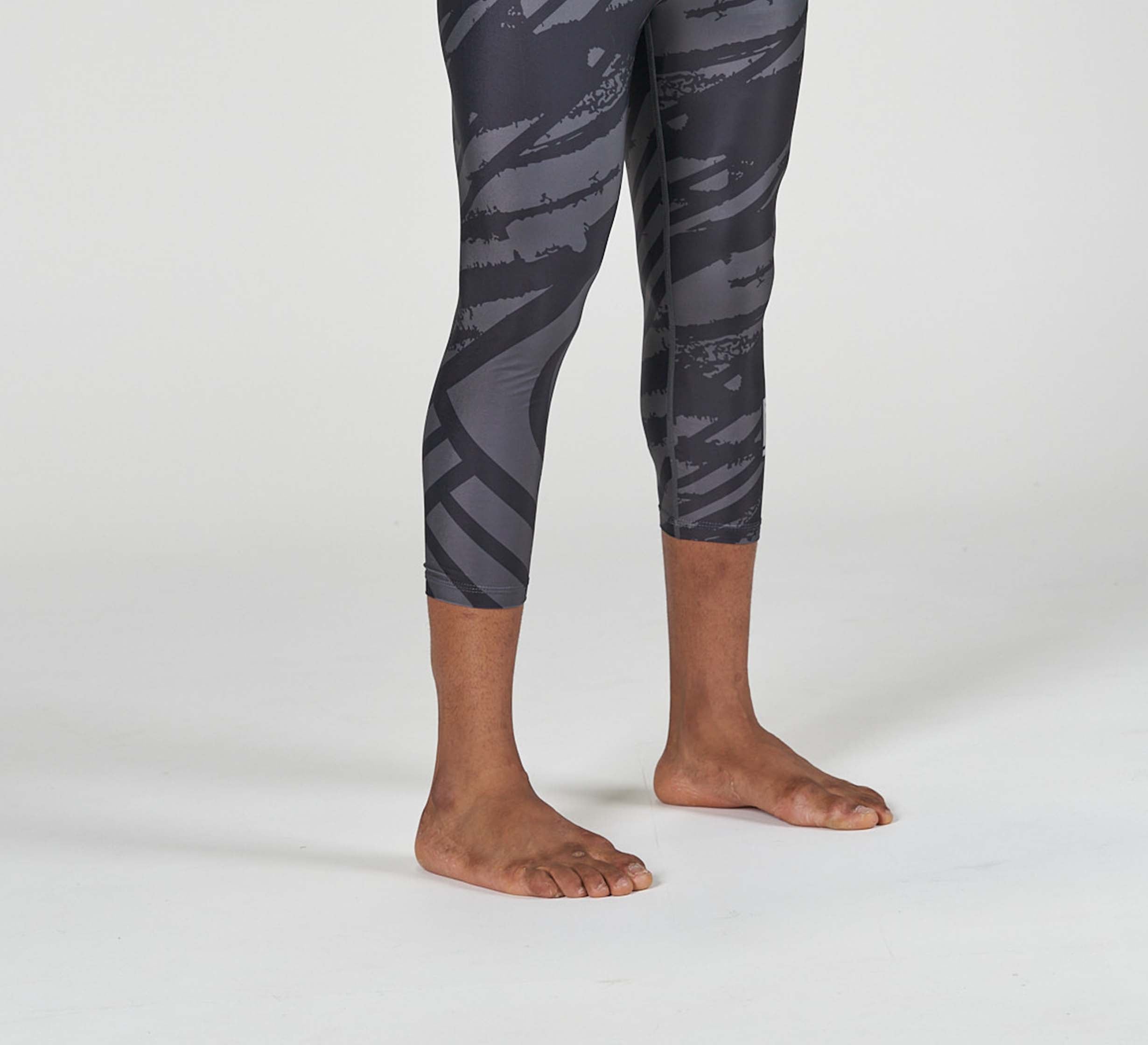 A person stands barefoot on a white surface, wearing Battle Flex Lite Spat Black leggings with a black and gray pattern. Only their legs and feet are visible.