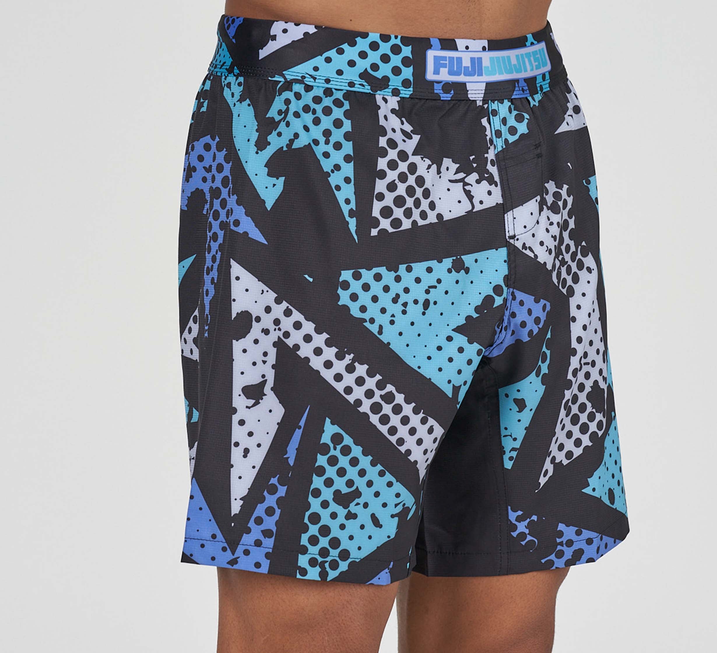 A person wearing Shuriken Flex Lite Shorts Blue/Purple, featuring a bold geometric pattern and Fuji Jiu Jitsu waistband. Only the lower torso and upper legs are visible.