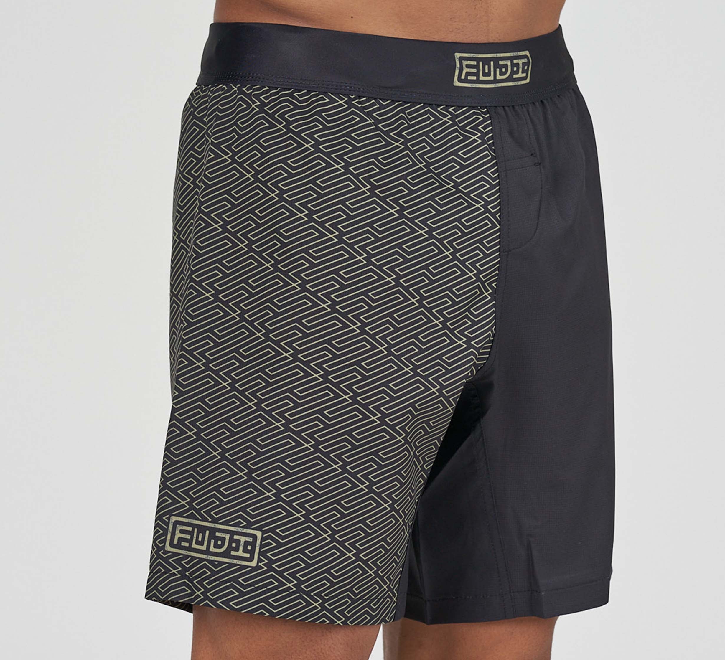 Man wearing Bushido Flex Lite Shorts featuring a black design with a geometric beige pattern on one side and logo patches on the waistband and leg.