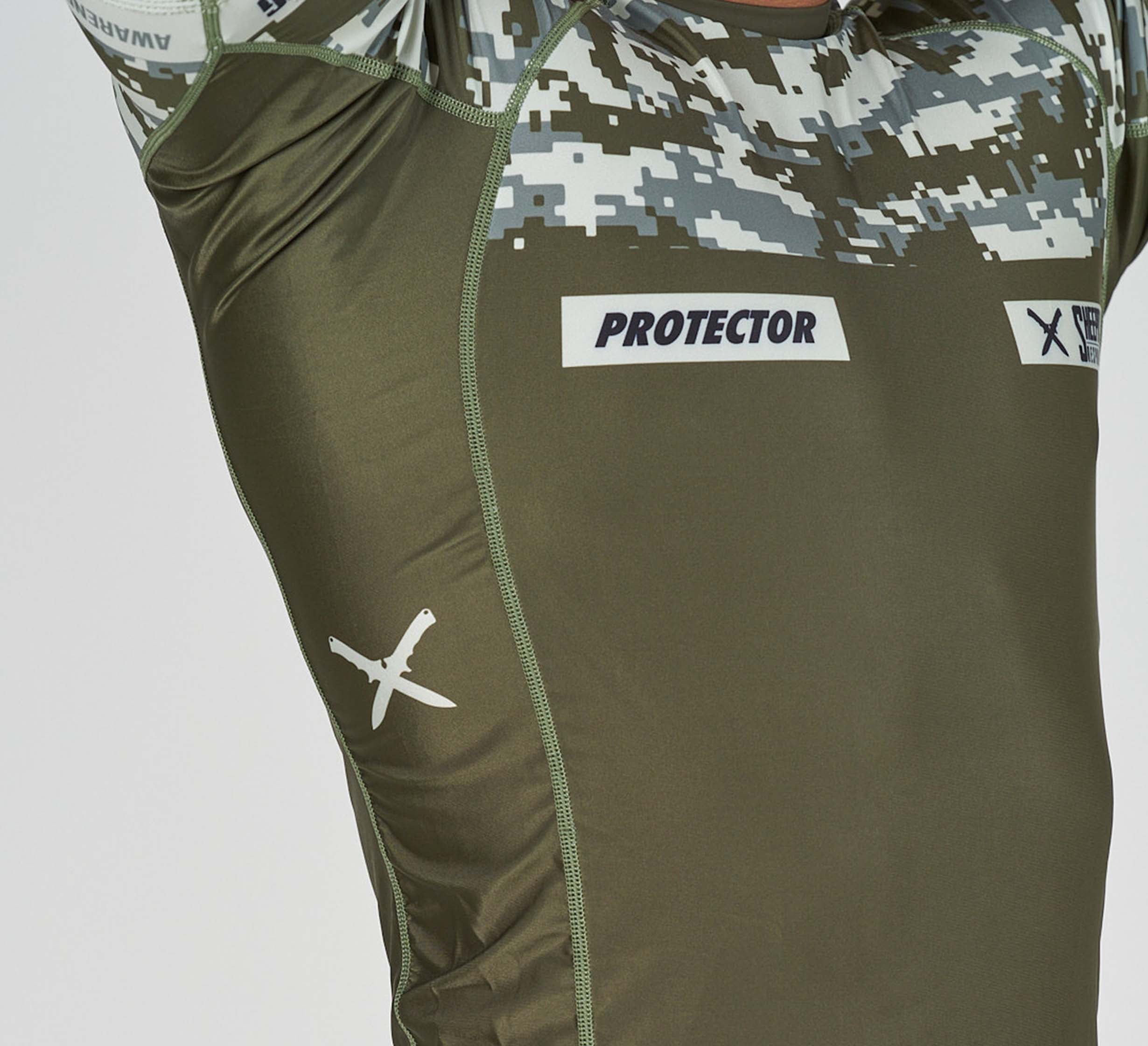 Close-up of a person wearing the Sheepdog Response Flex Lite Rashguard in Military Green, featuring camouflage details, "PROTECTOR" text, and a white X-shaped logo on the chest and side.