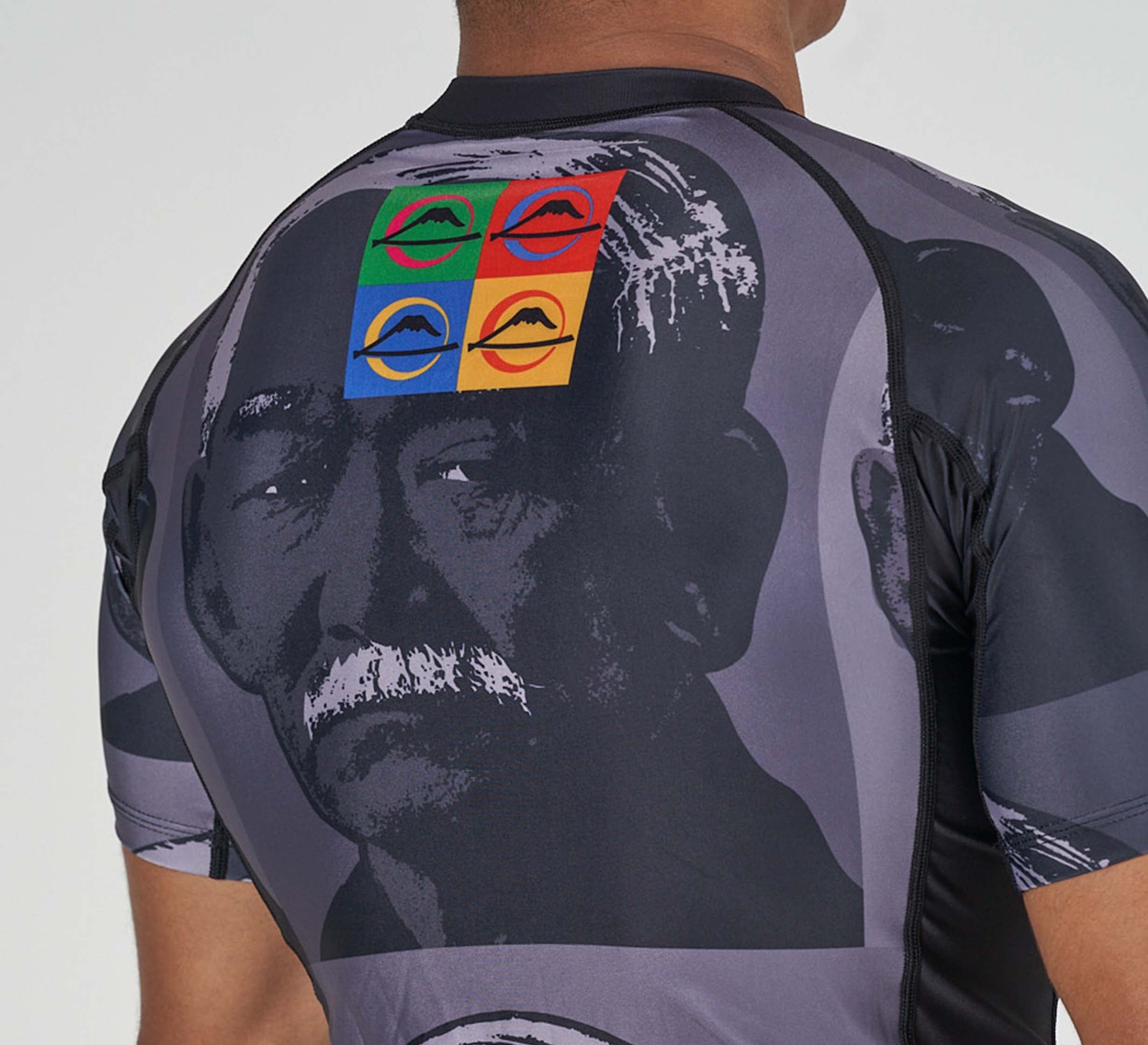 A person wearing the Kano Flex Lite Rashguard, which features a grayscale portrait of an older man with a mustache on the front and a colorful pop-art patch of four faces in red, green, blue, and yellow squares on the upper back.