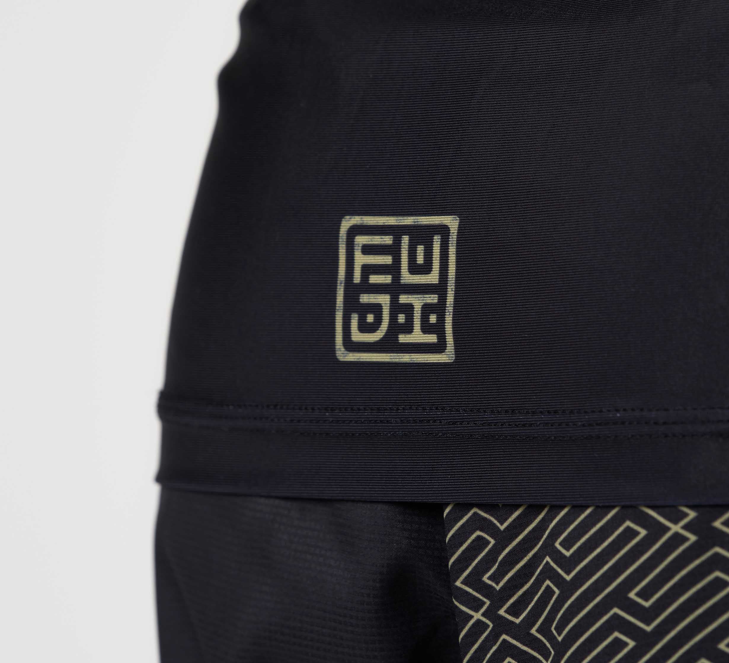 Close-up of the Bushido Flex Lite Rashguard in black, featuring a gold geometric logo made of stylized square letters and a matching gold maze-like pattern on the fabric.