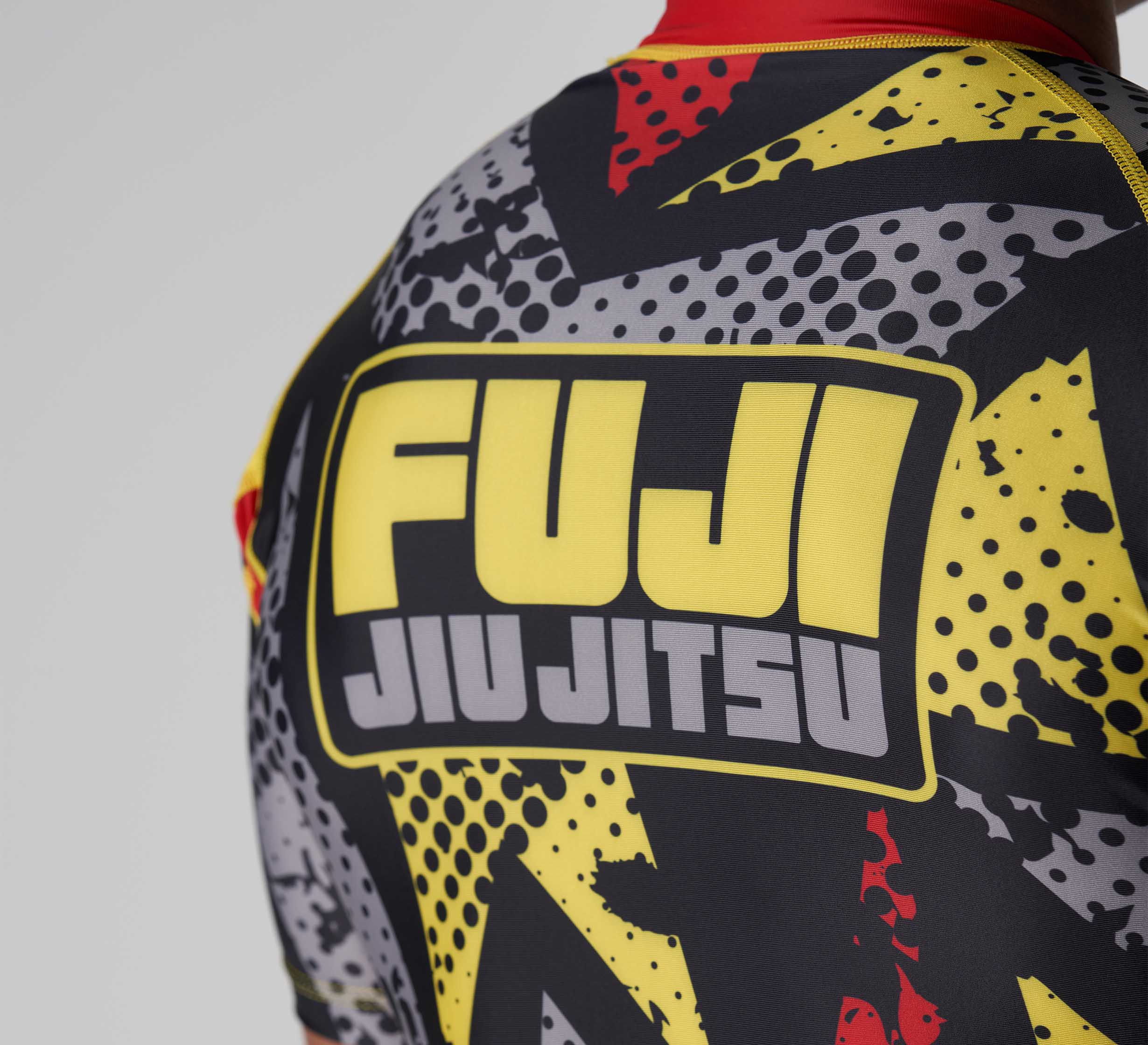 A close-up of the back of a person wearing the Shuriken Flex Lite Rashguard Black/Yellow, featuring bold FUJI JIU JITSU text in yellow and white over a black, yellow, and gray graphic design.