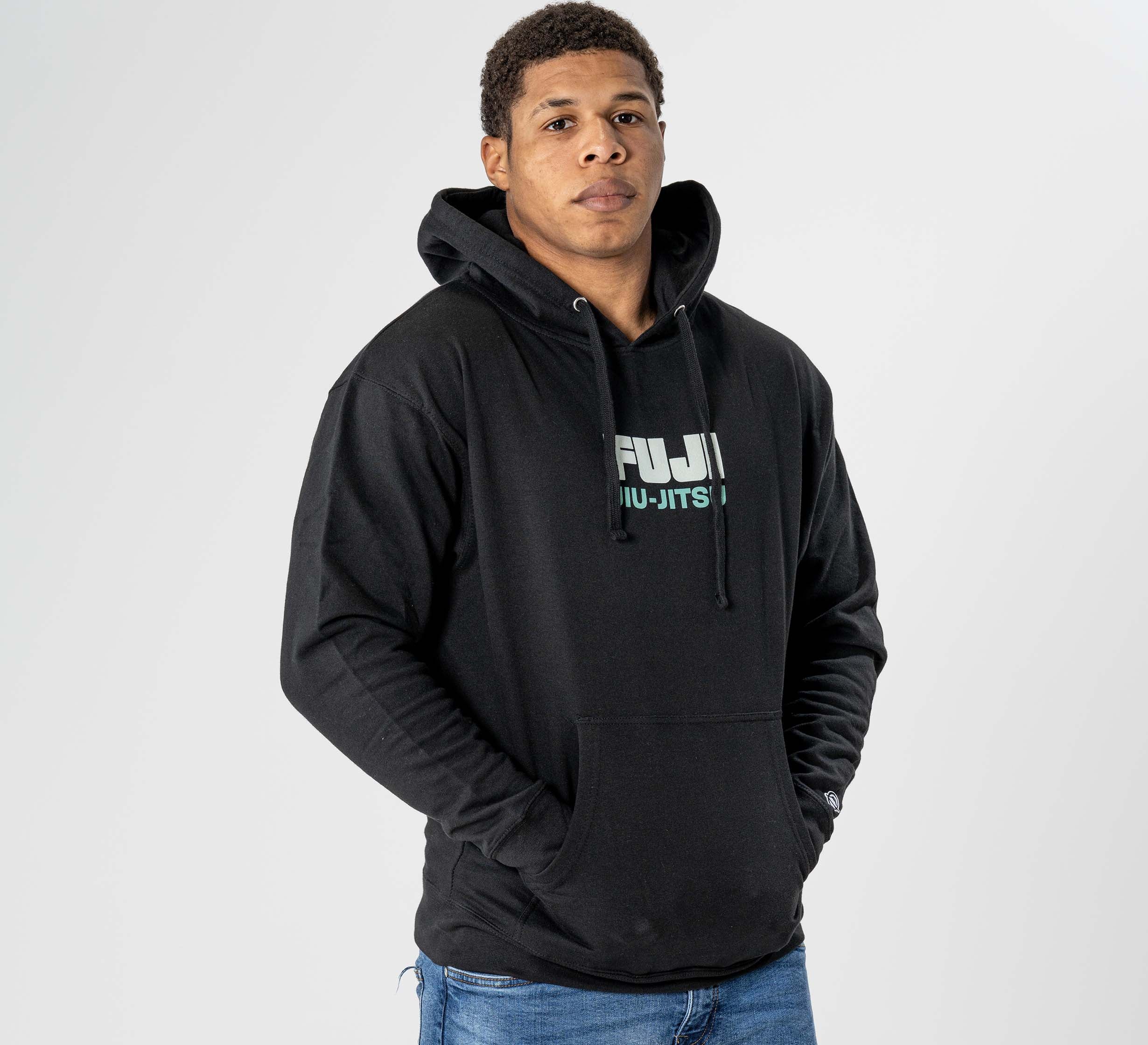 A person stands against a plain white background, hands in the pocket of the Black Belt Mentality Hoodie Black, featuring “FUJI JIU-JITSU” on the front, and wears a neutral expression.