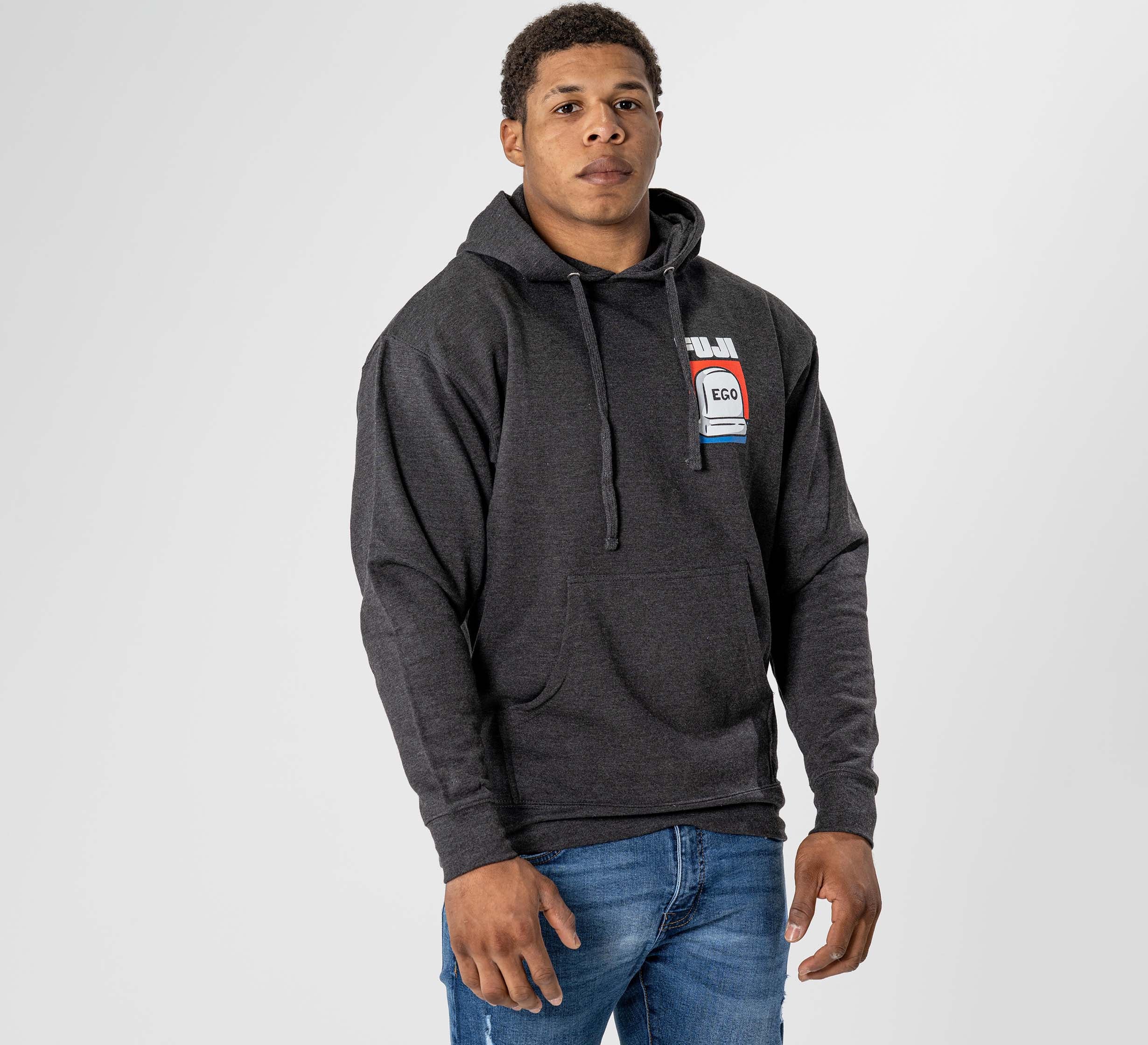 A man stands against a plain light background wearing the Submit Your Ego Hoodie in Heather Black, featuring a chest graphic name tag that reads "Hello, my name is Ego," paired with blue jeans.