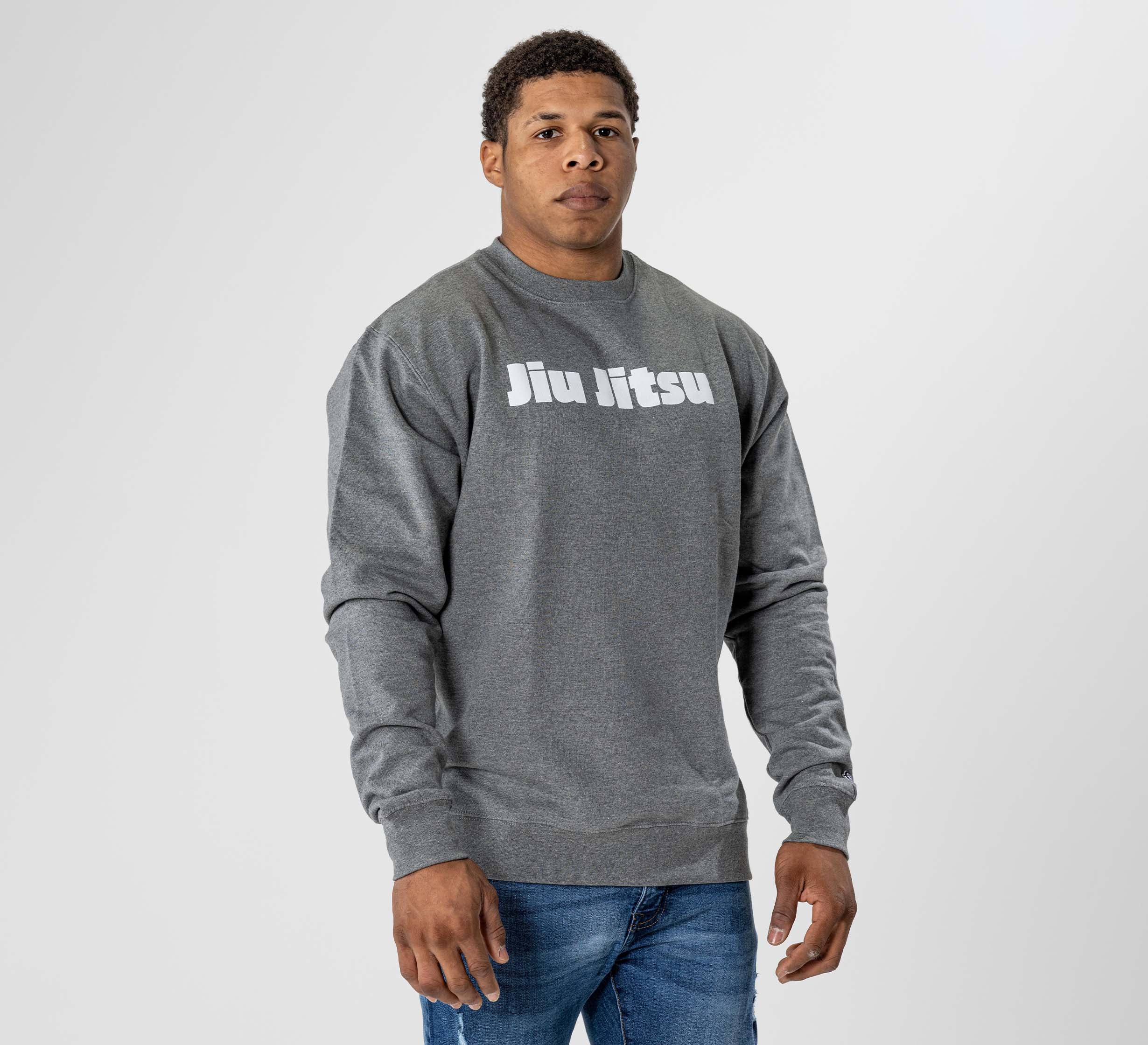 A man in the Jiu Jitsu Player Crewneck Heather Grey, featuring "Jiu Jitsu" in white on the chest, stands against a light gray background. He has short curly hair and is wearing blue jeans.