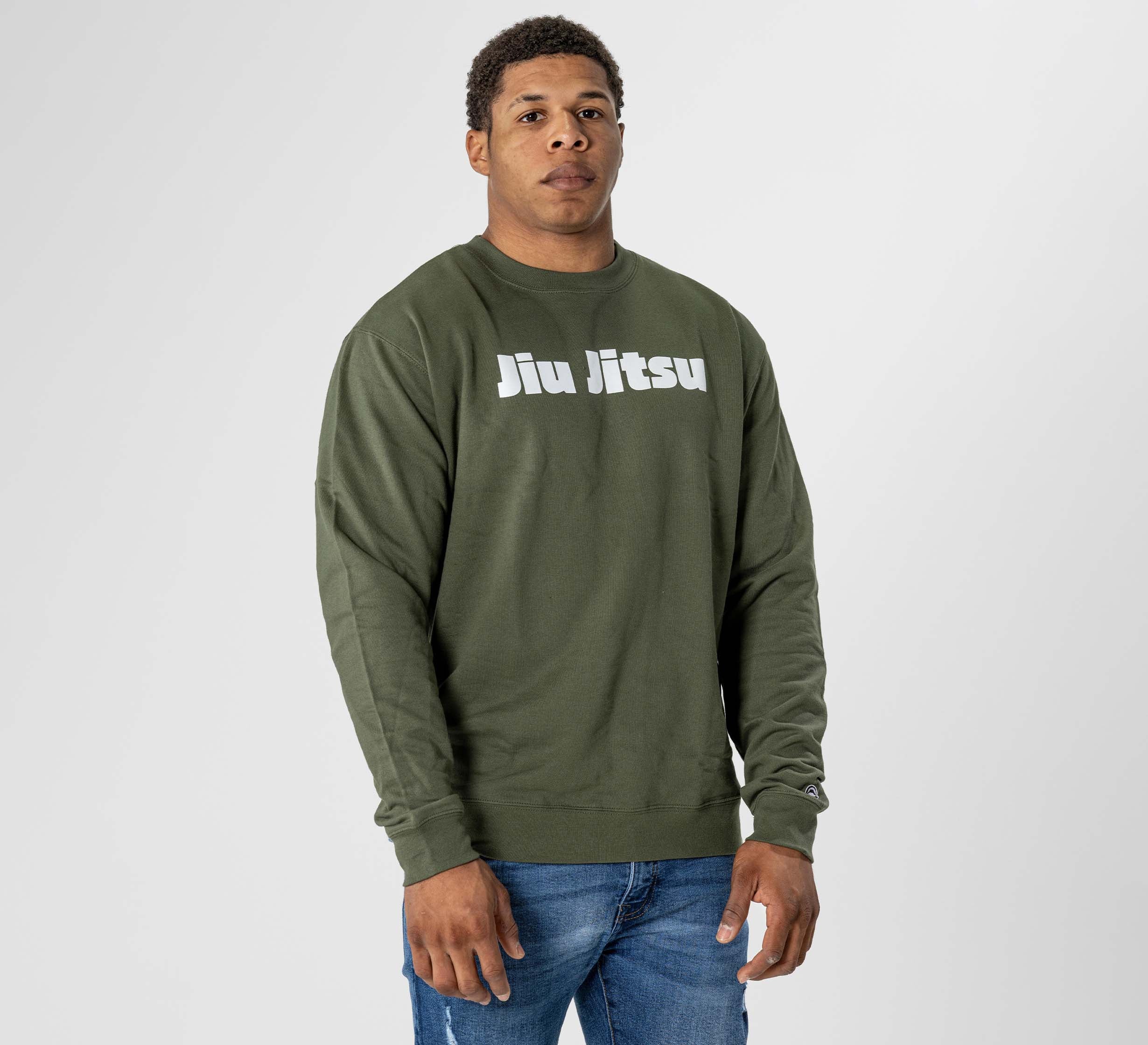 A man stands against a plain light background, wearing the Jiu Jitsu Player Crewneck in military green and blue jeans. "Jiu Jitsu" is printed in white on the chest. He looks directly at the camera with a neutral expression.