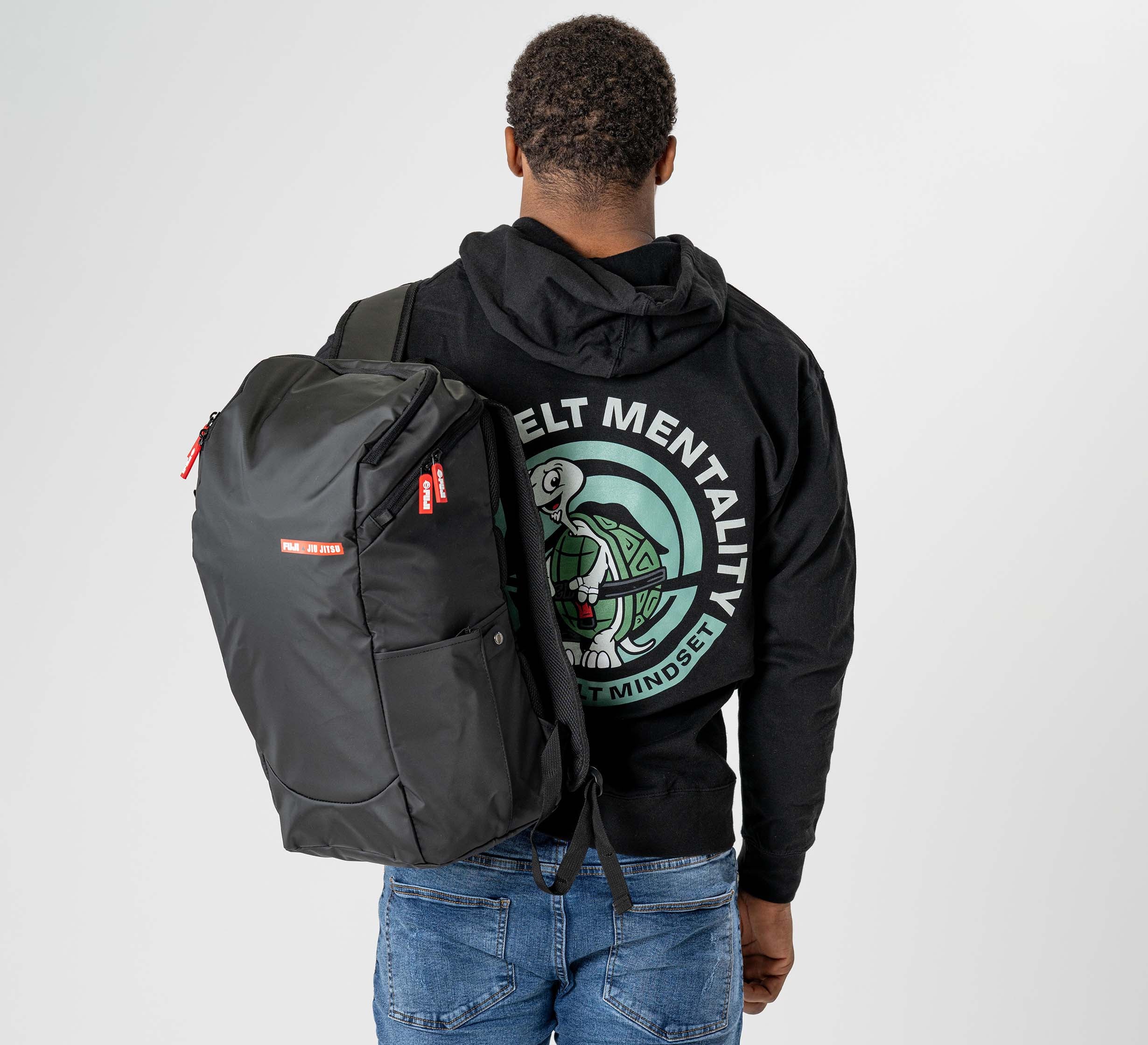 A person in a black graphic hoodie and blue jeans faces away against a plain background, carrying the Urban Day Backpack Black over one shoulder.
