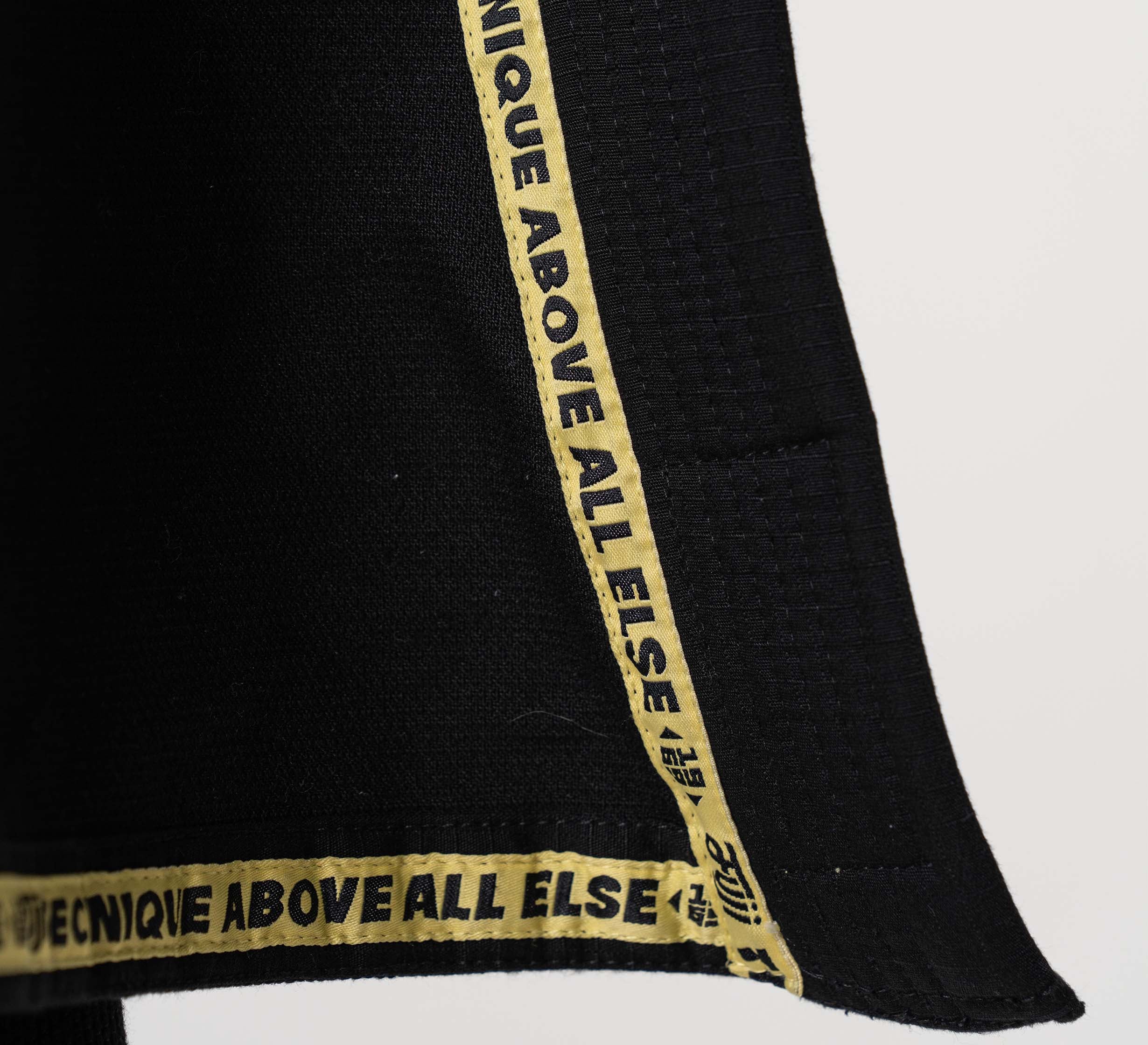 A close-up of the Flow-Tech BJJ Gi Black/Gold shows its black fabric edge with a gold trim featuring bold black "TECHNIQUE ABOVE ALL ELSE" text. The background is plain white.