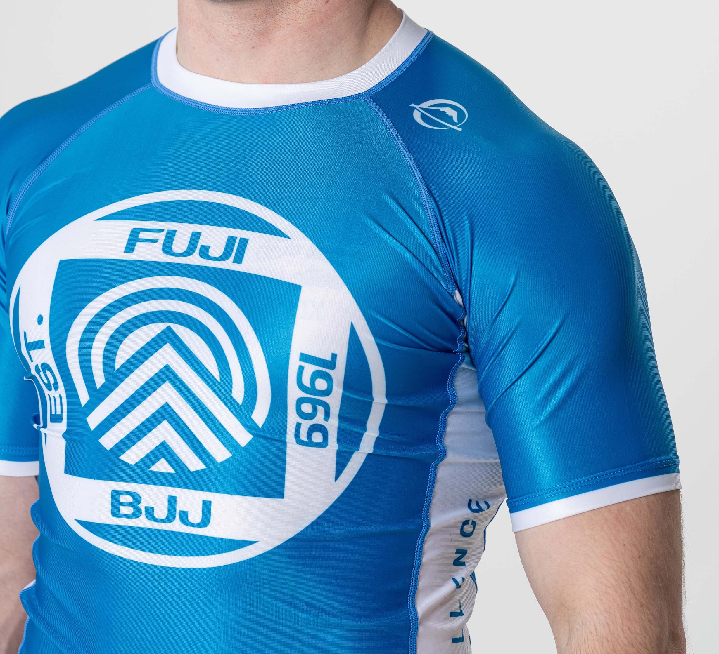 A person wears the BJJ Flex Lite Rashguard Blue, featuring a fitted design with blue and white colors, a large circular logo on the chest, and a smaller shoulder logo, standing against a plain light background.