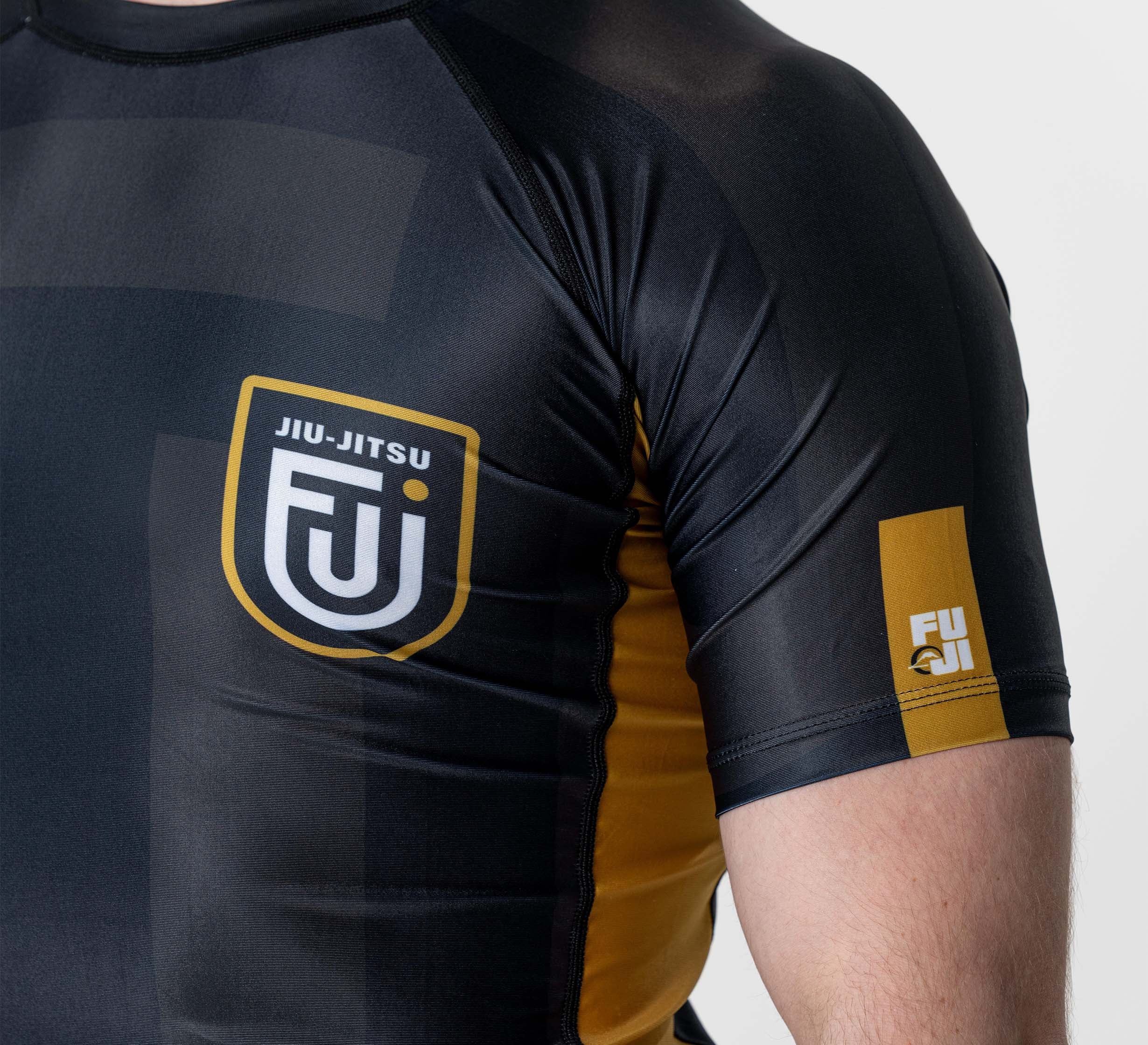 A person wearing a Rebel Flex Lite Rashguard Black, featuring black and yellow colors, the FUJI logo and “JIU-JITSU” on the chest, plus another FUJI logo on the left sleeve.