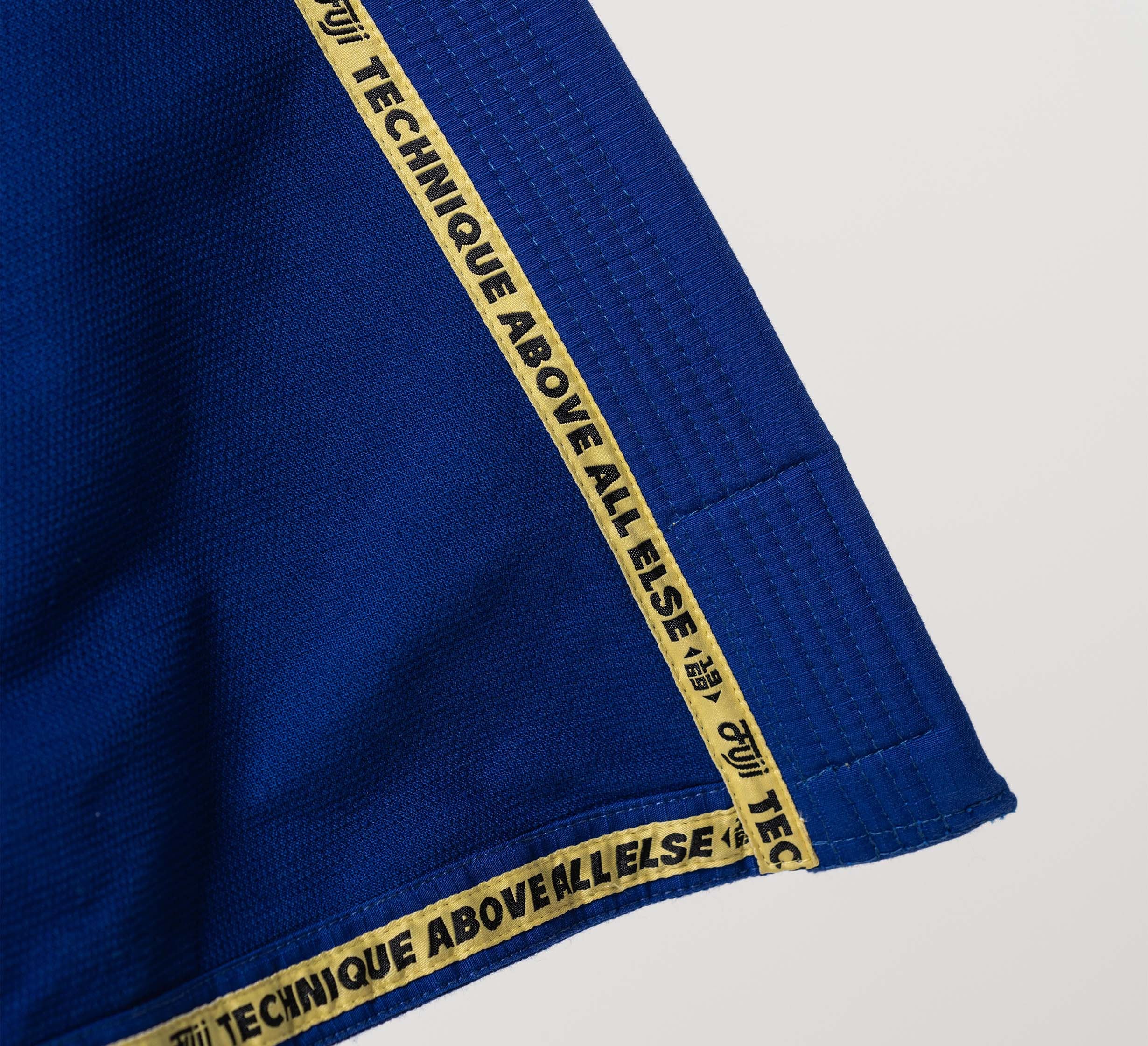 A close-up of the Flow-Tech BJJ Gi Blue/Gold shows its blue fabric edge with a yellow trim featuring “TECHNIQUE ABOVE ALL ELSE” in black, repeated along the border.