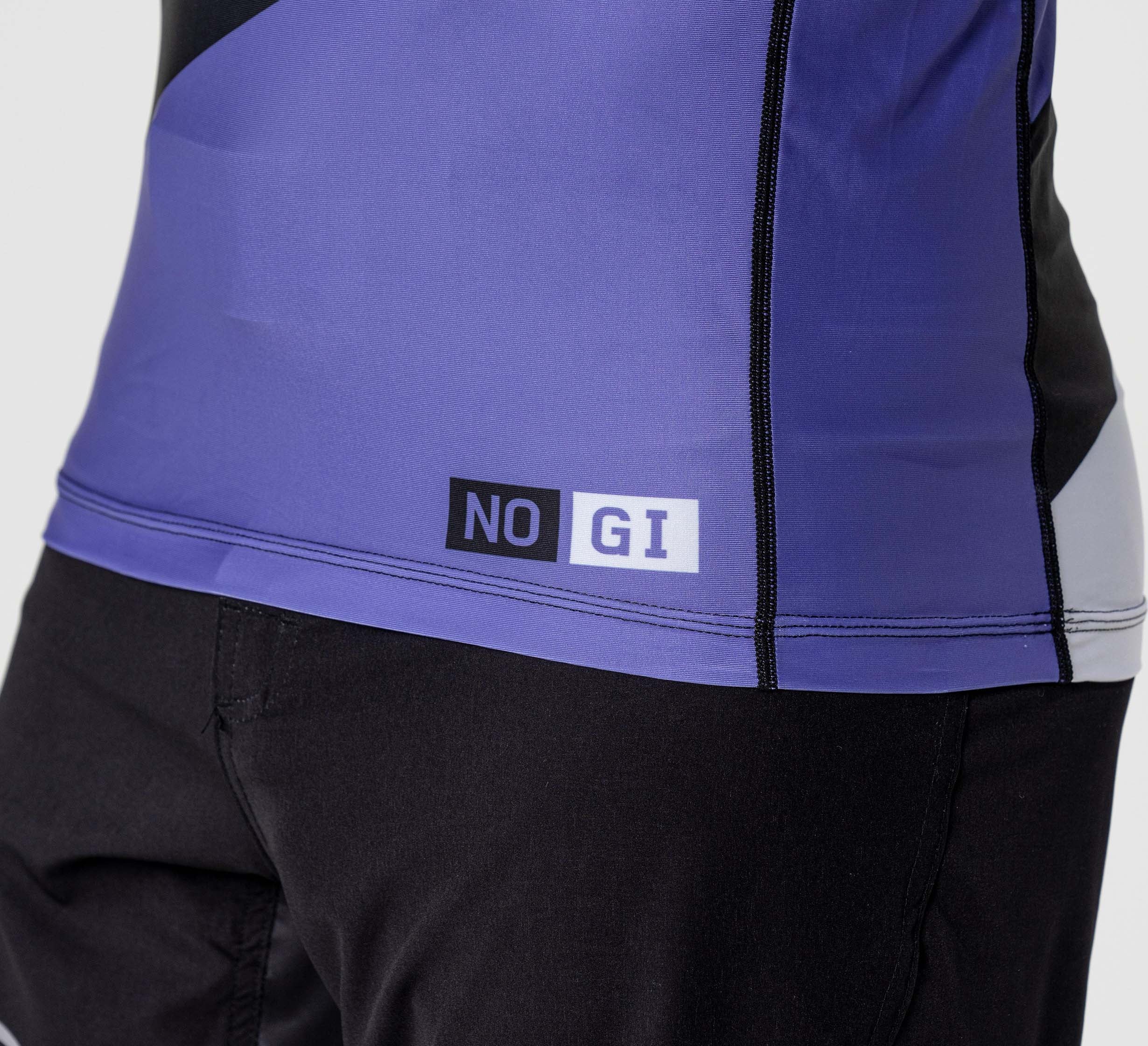 A close-up of a person wearing the Pacer Flex Lite Long Sleeve Rashguard Purple, featuring a NO GI label near the hem, paired with black shorts.
