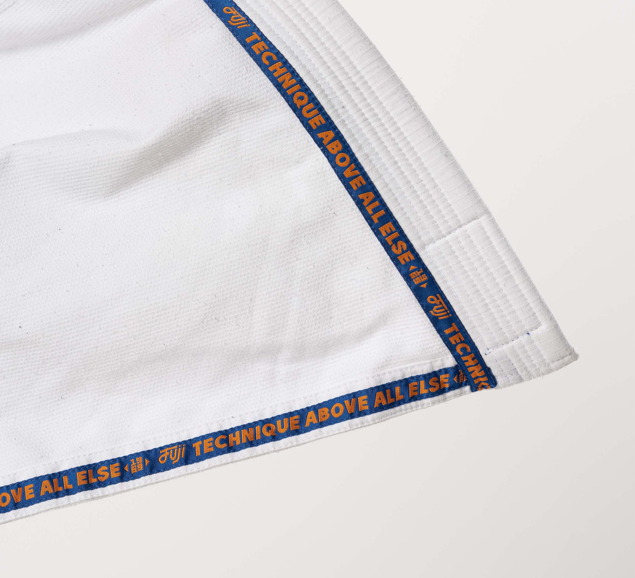 Close-up of the Flow-Tech BJJ Gi in White/Navy/Orange, featuring "TECHNIQUE ABOVE ALL ELSE" along the edge, with visible fabric texture and detailed stitching.