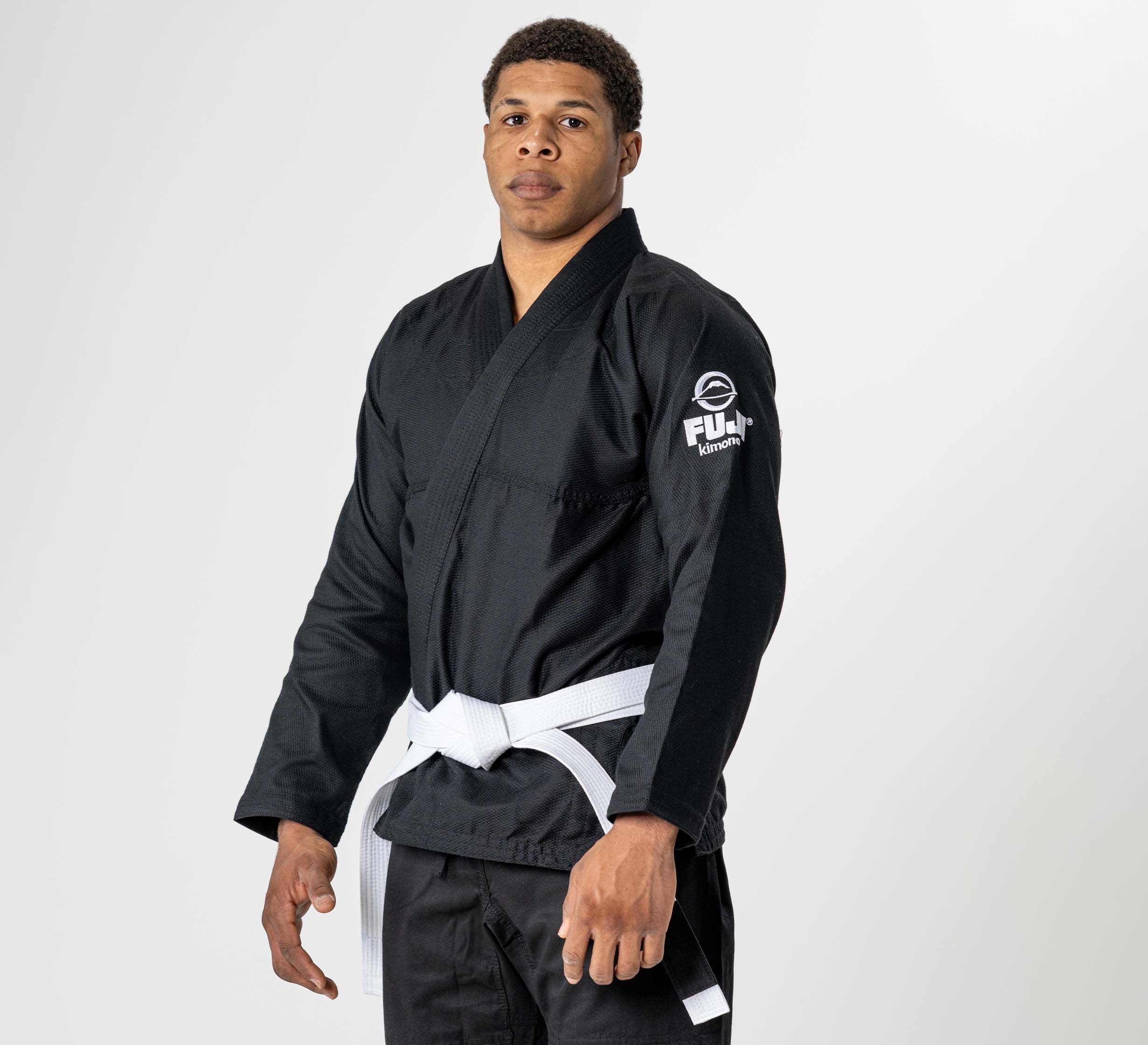 A man stands against a plain white background wearing the Starter Adult BJJ Gi Black with a white belt. The gi features a FUJI logo on the sleeve. He looks directly at the camera with a serious expression.