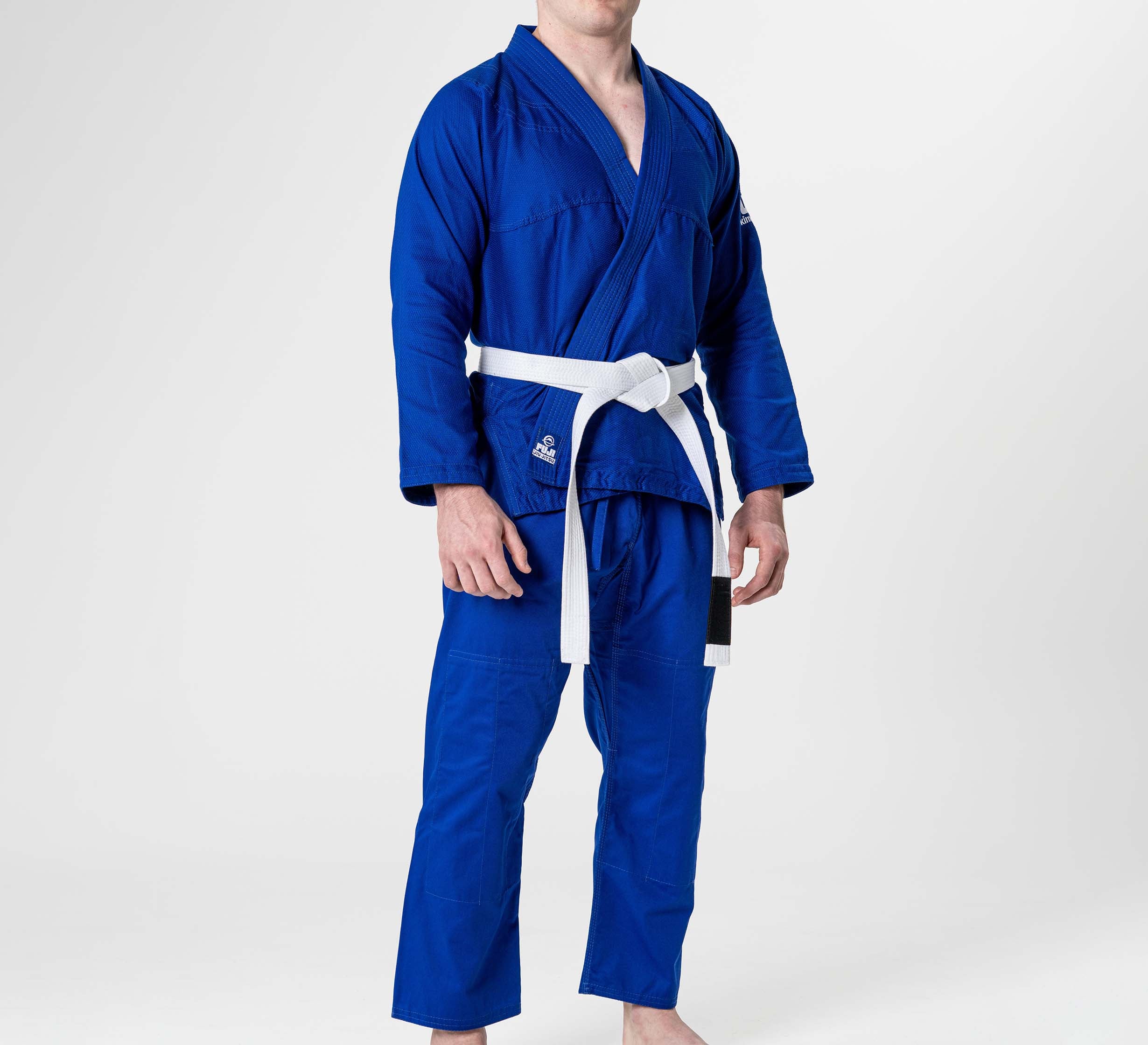A person wears the Starter Adult BJJ Gi Blue with a white belt, barefoot, against a plain white background. The gi features a V-neck and long sleeves; the person's face is partially out of frame.