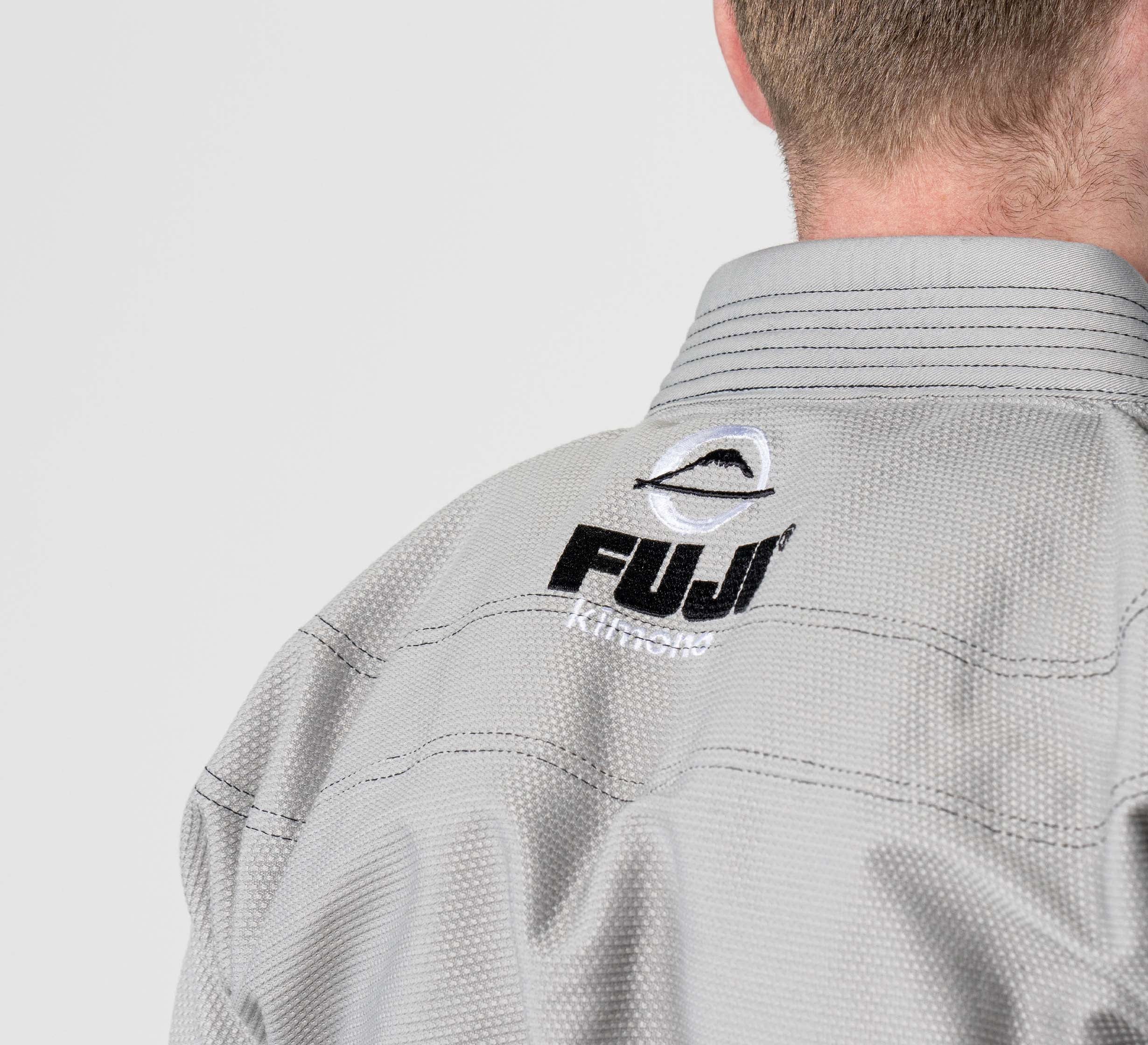 Close-up of a person wearing the Lightweight BJJ Gi Grey with FUJI Kimonos and a black and white mountain logo embroidered on the back of the collar. The background is plain and light-colored.