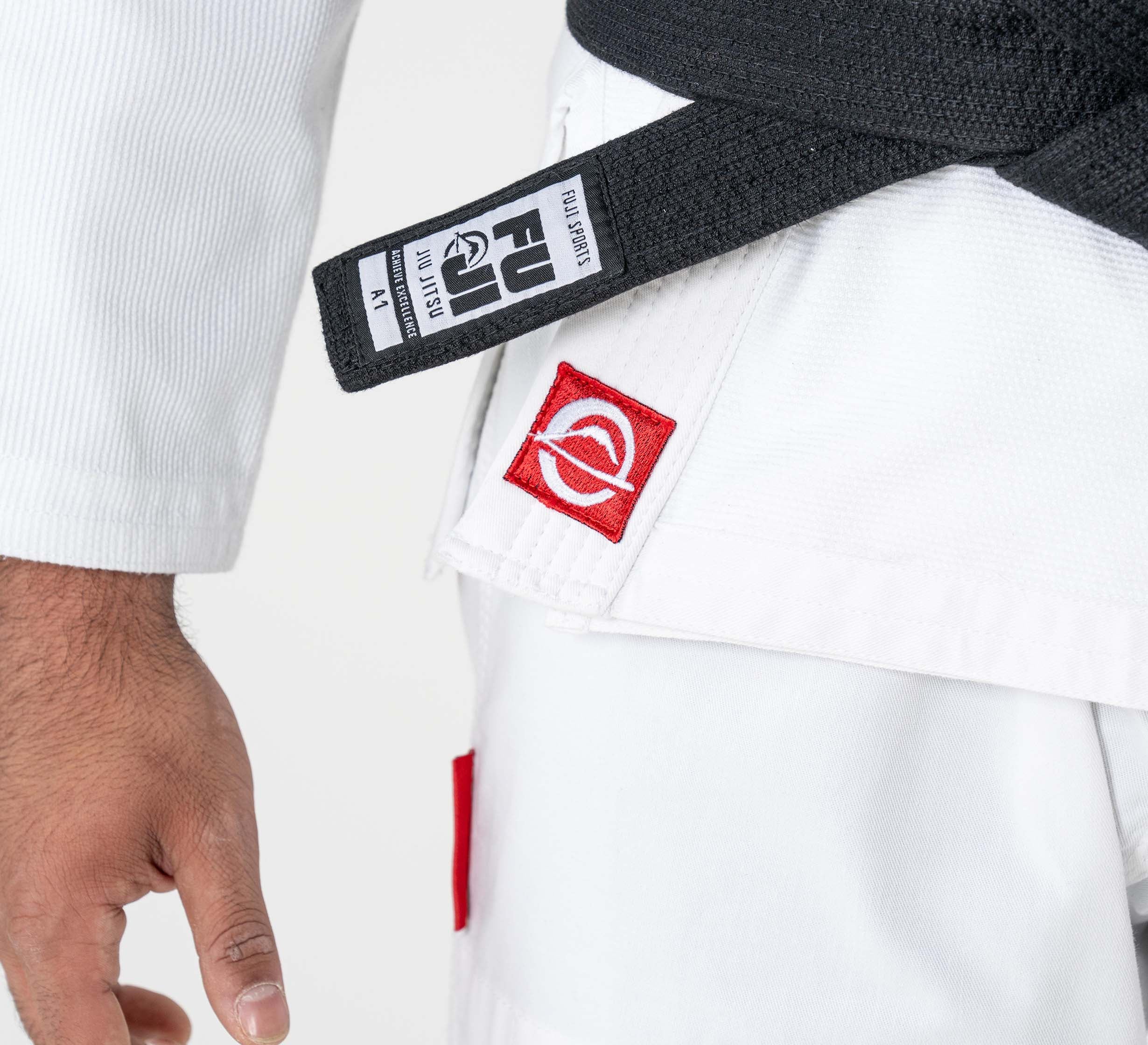 A person wearing the Kids Ultra Lite 300 BJJ Gi in white with a black belt, featuring red patches and showing part of their left arm and hand.
