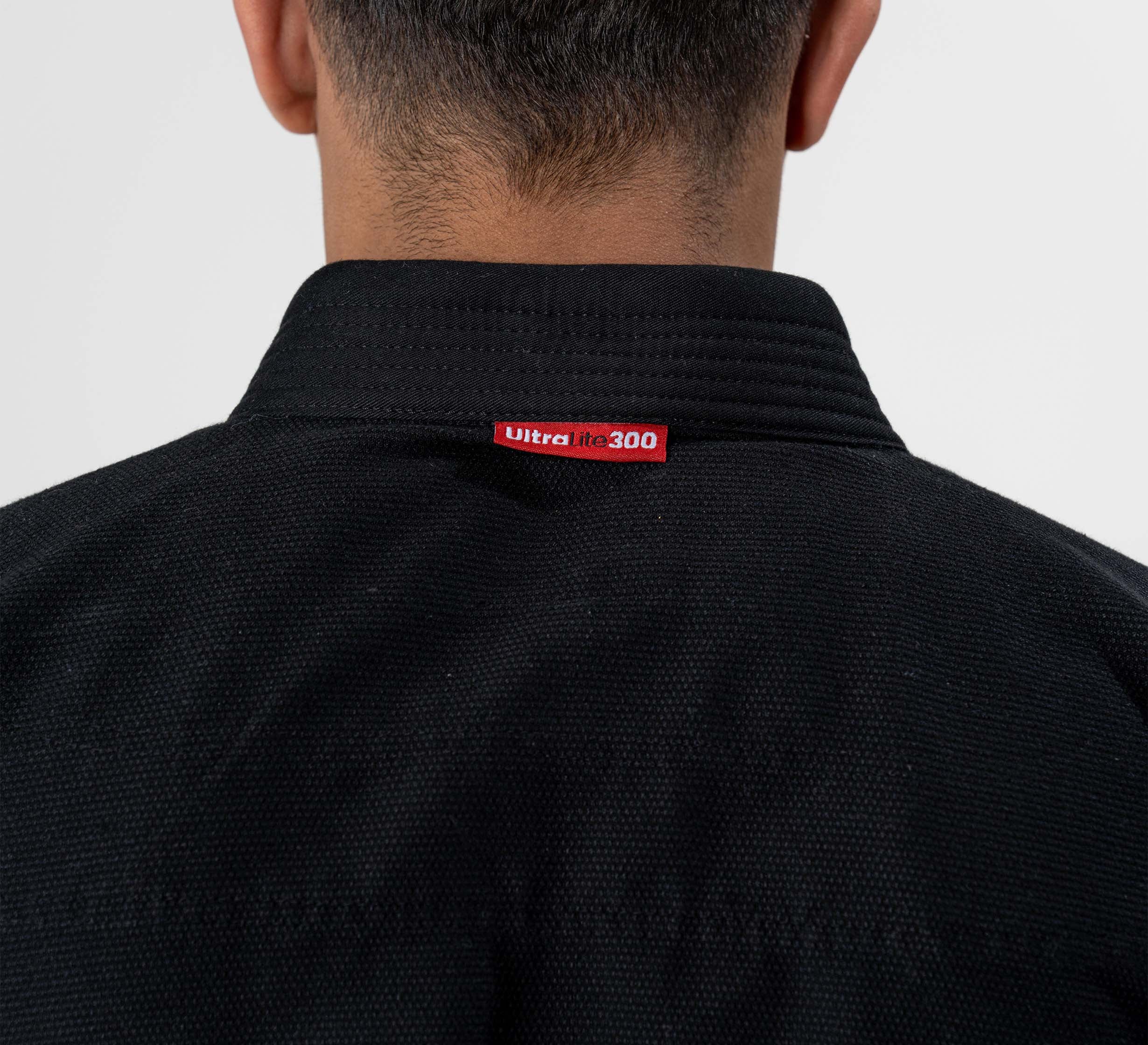 A person with a short haircut is seen from behind wearing the Ultra Lite 300 BJJ Gi in black, with the red and white Ultra Lite 300 label visible on the back of the collar.