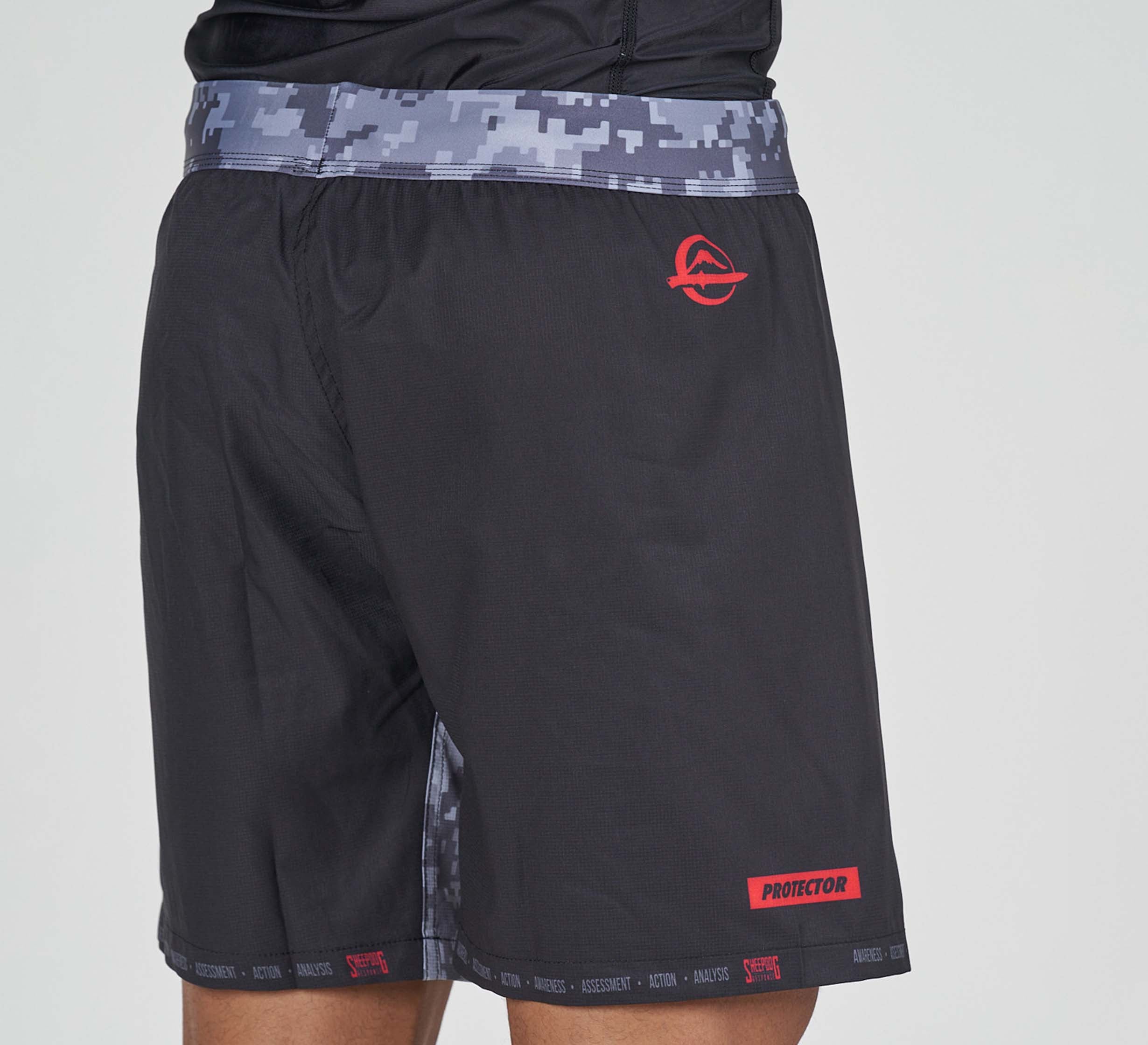 A person wears Sheepdog Response Flex Lite Shorts Black featuring a gray digital camo waistband, red logo on the left hip, and "PROTECTOR" in red on the lower left leg. The upper body is partially visible.