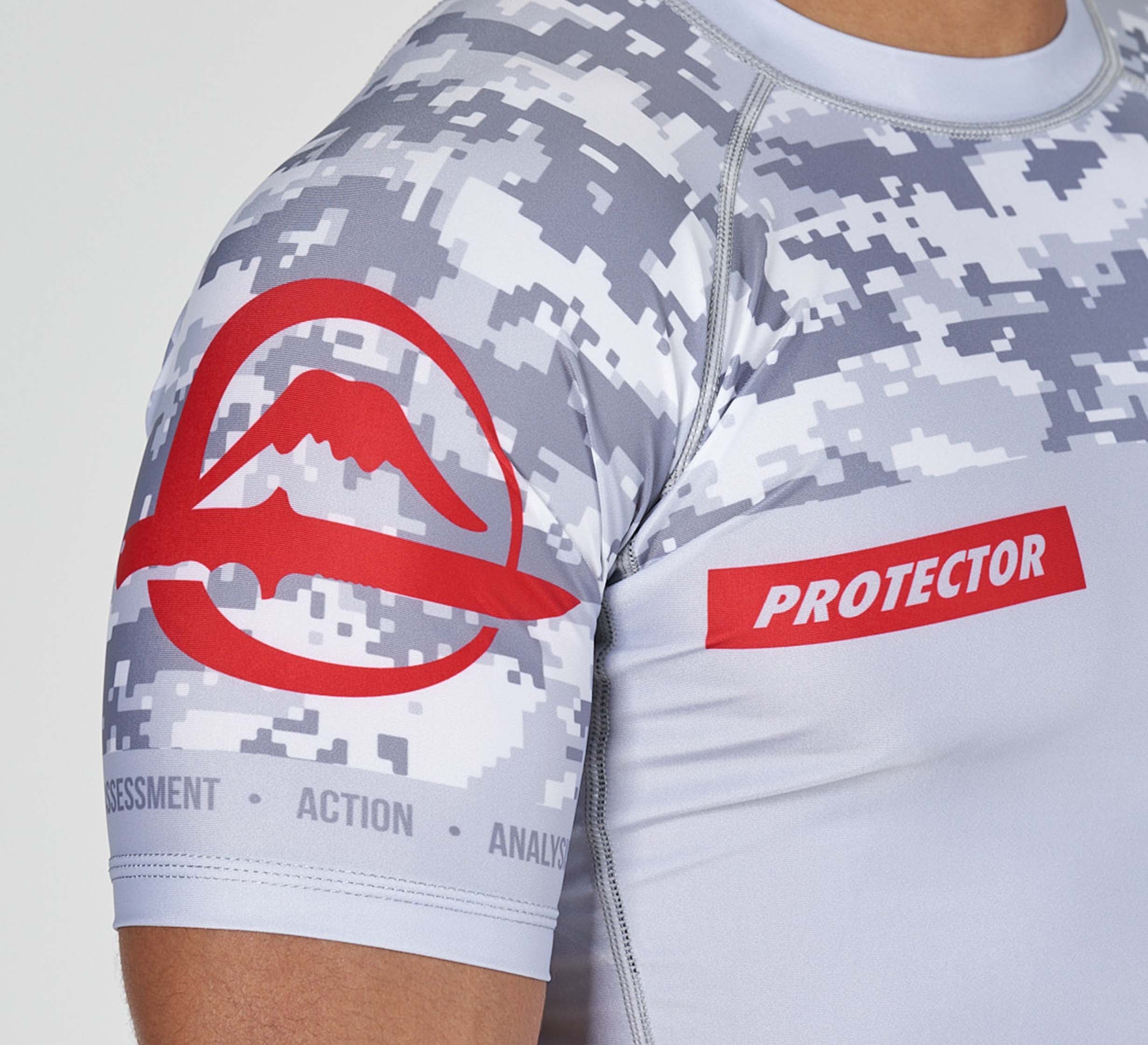 A close-up of a person wearing the Sheepdog Response Flex Lite Rashguard Grey, featuring white and gray digital camouflage, a red PROTECTOR label, a red mountain logo on the sleeve, and “ASSESSMENT ACTION ANALYSIS” on the lower sleeve.