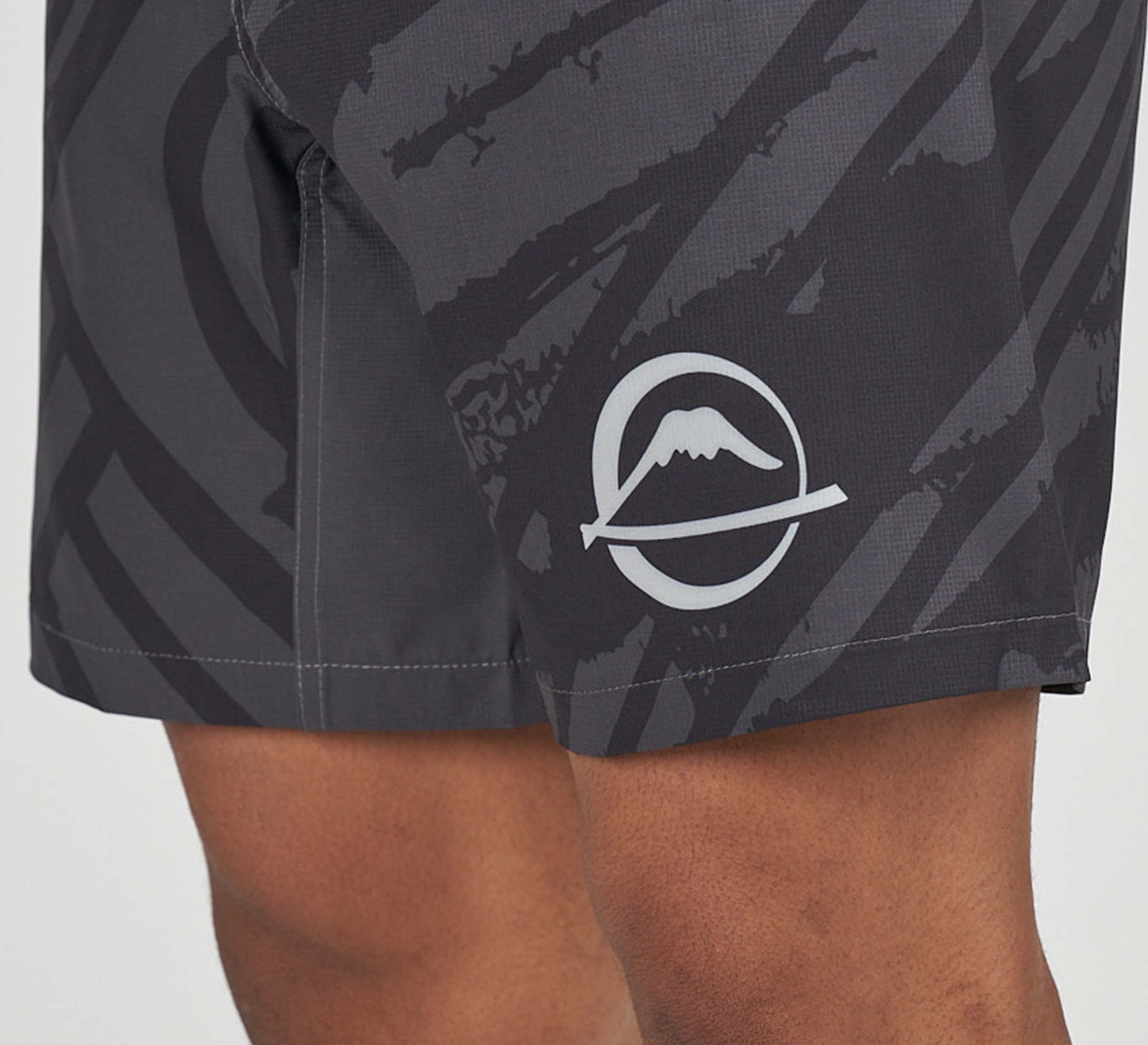 Close-up of a person's leg wearing Battle Flex Lite Shorts Black with a white circular mountain logo on the side. The shorts feature an abstract geometric pattern in various shades of gray.