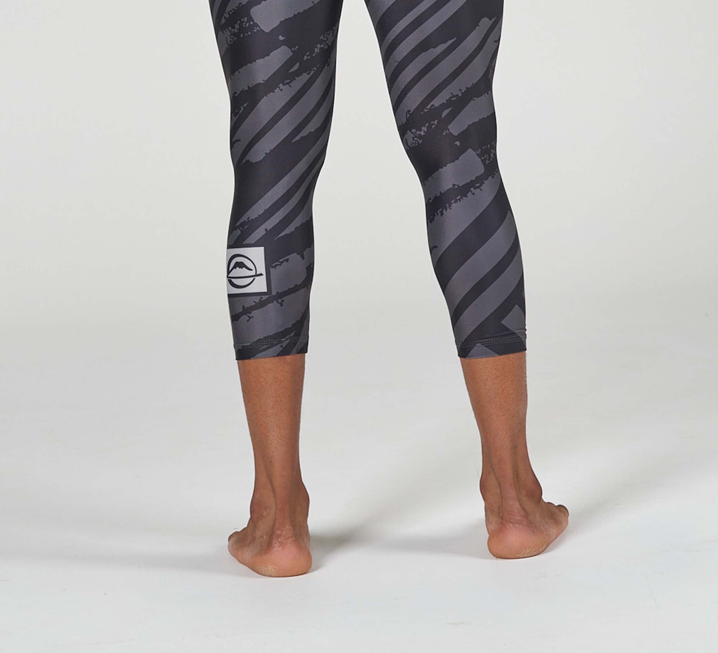A person stands barefoot on a light surface, shown from the calves down, wearing Battle Flex Lite Spat Black leggings featuring a black and gray pattern with a small white logo on the left leg.