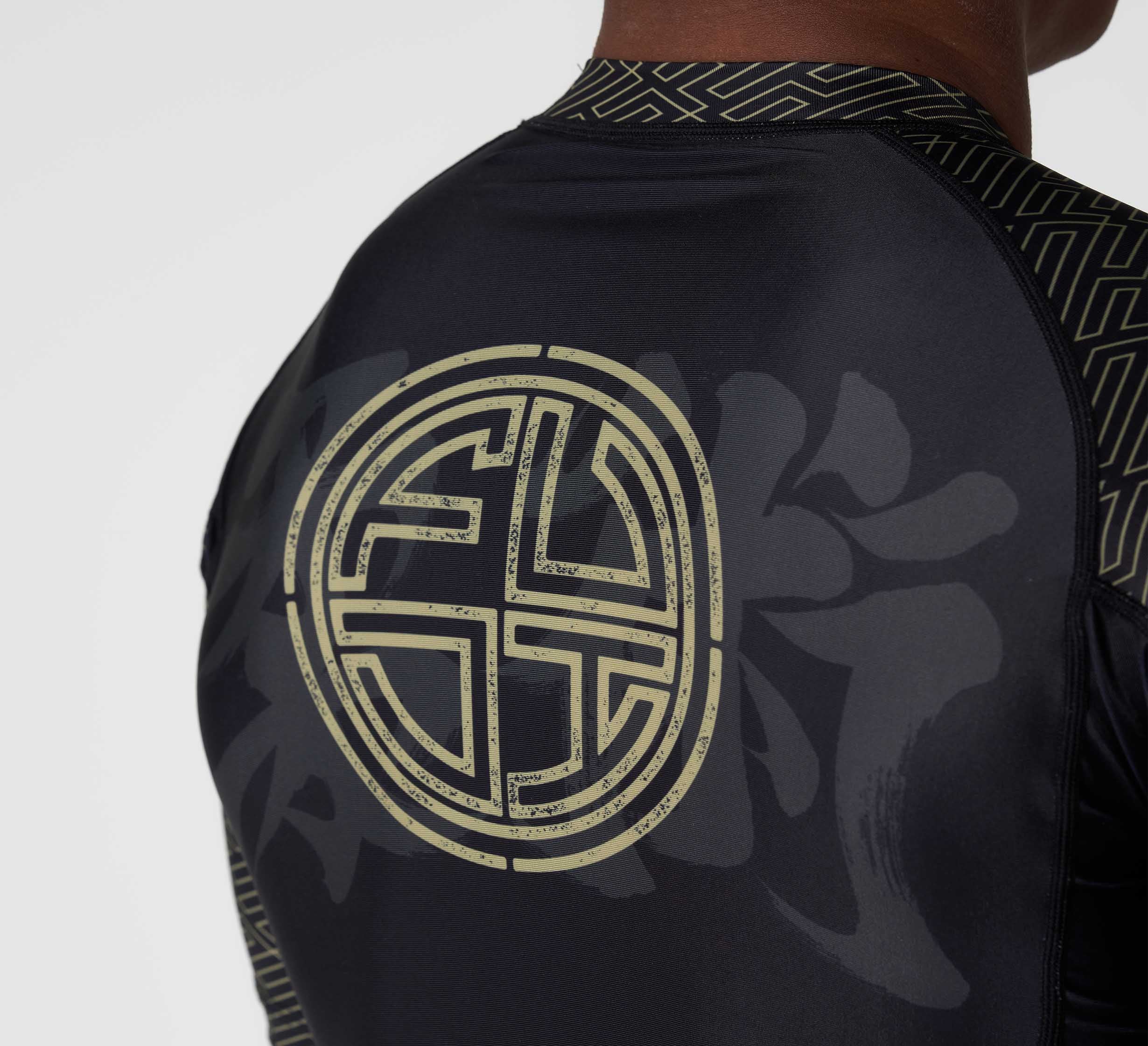 Close-up of the back of a person wearing the Bushido Flex Lite Rashguard, a black shirt with a large circular gold geometric design, subtle dark background patterns, and patterned collar and sleeves.