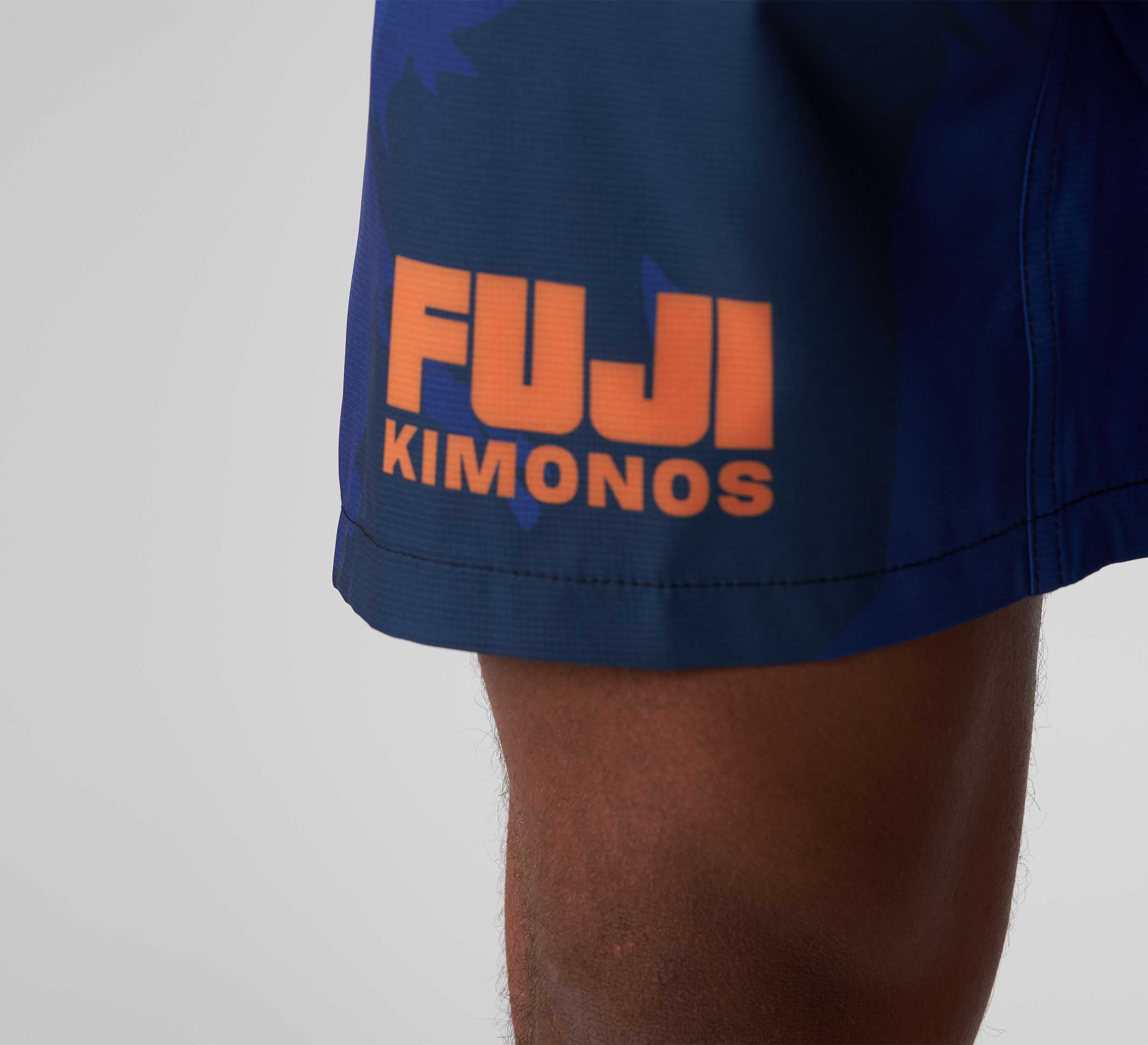 Close-up of a person's thigh wearing Hanzo Flex Lite Shorts Blue, featuring "FUJI KIMONOS" in bold orange letters near the hem. The background is plain and light-colored.