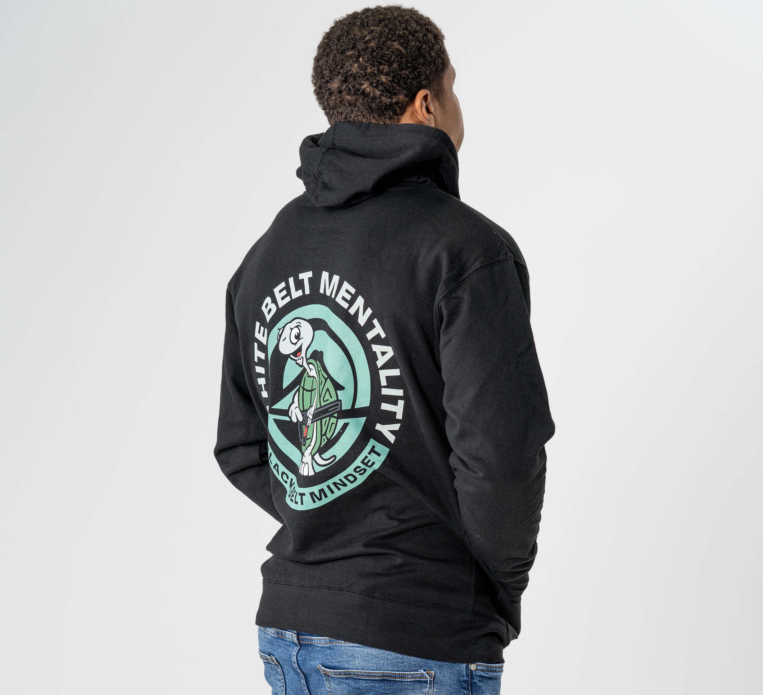 A person wearing the Black Belt Mentality Hoodie Black, featuring a large circular back graphic with "White Belt Mentality" and "Reset Mindset," plus a white belt martial artist illustration, stands with their back to the camera.