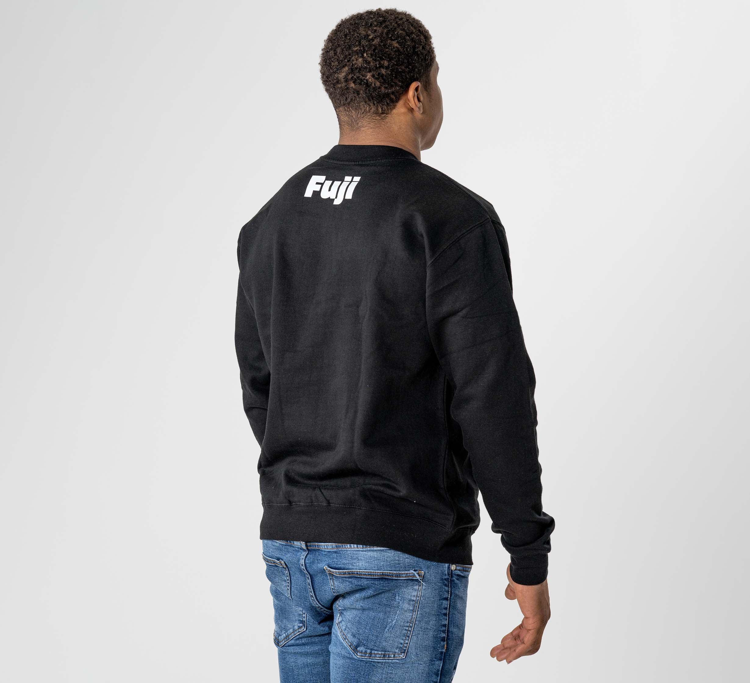 A person wears the Jiu Jitsu Player Crewneck Black, featuring "Fuji" in white on the back, styled with blue jeans against a plain light gray background.