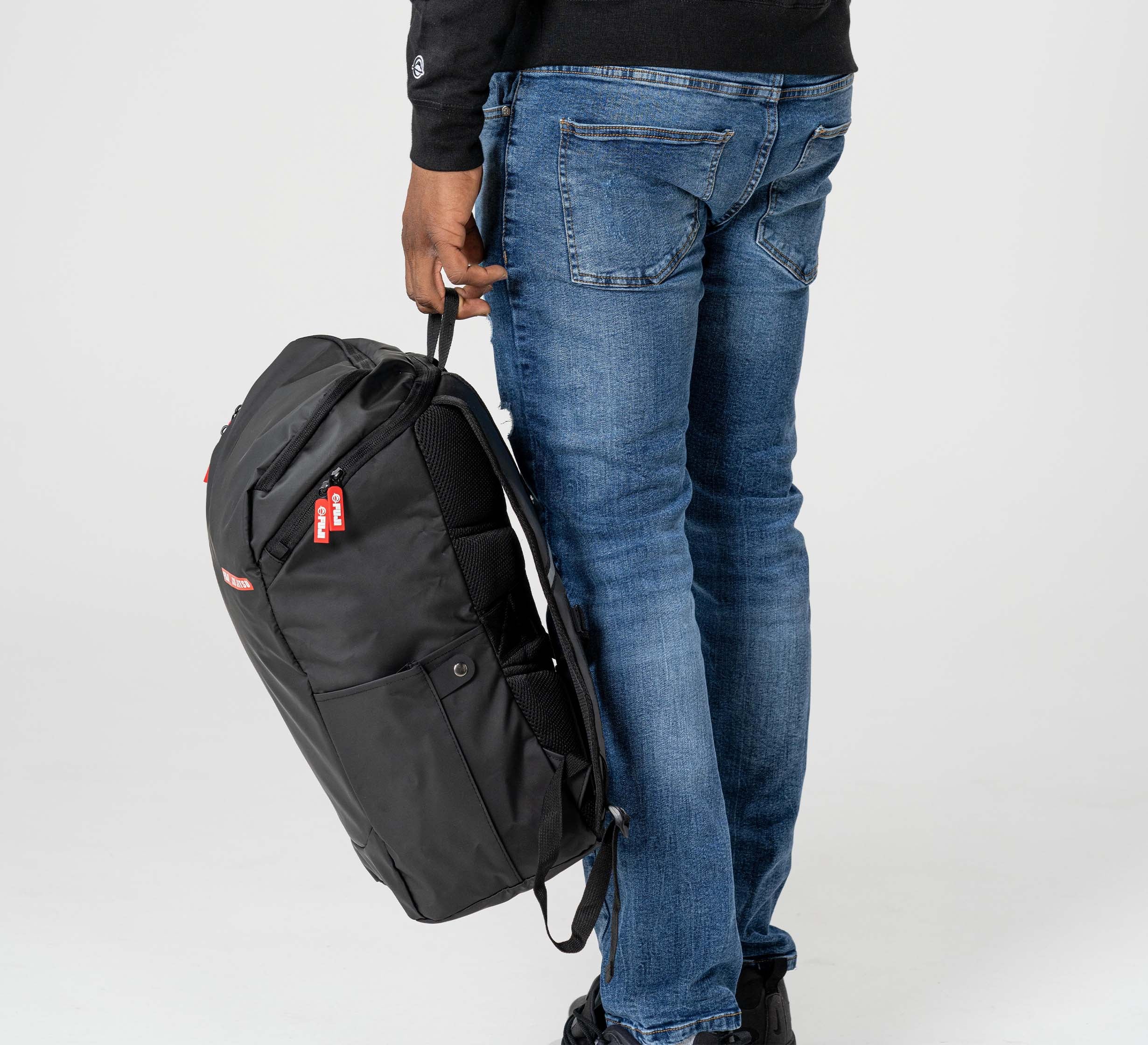A person in blue jeans and a black sweater stands holding the Urban Day Backpack Black by its side handle, featuring a red and white tag, against a plain light gray background.