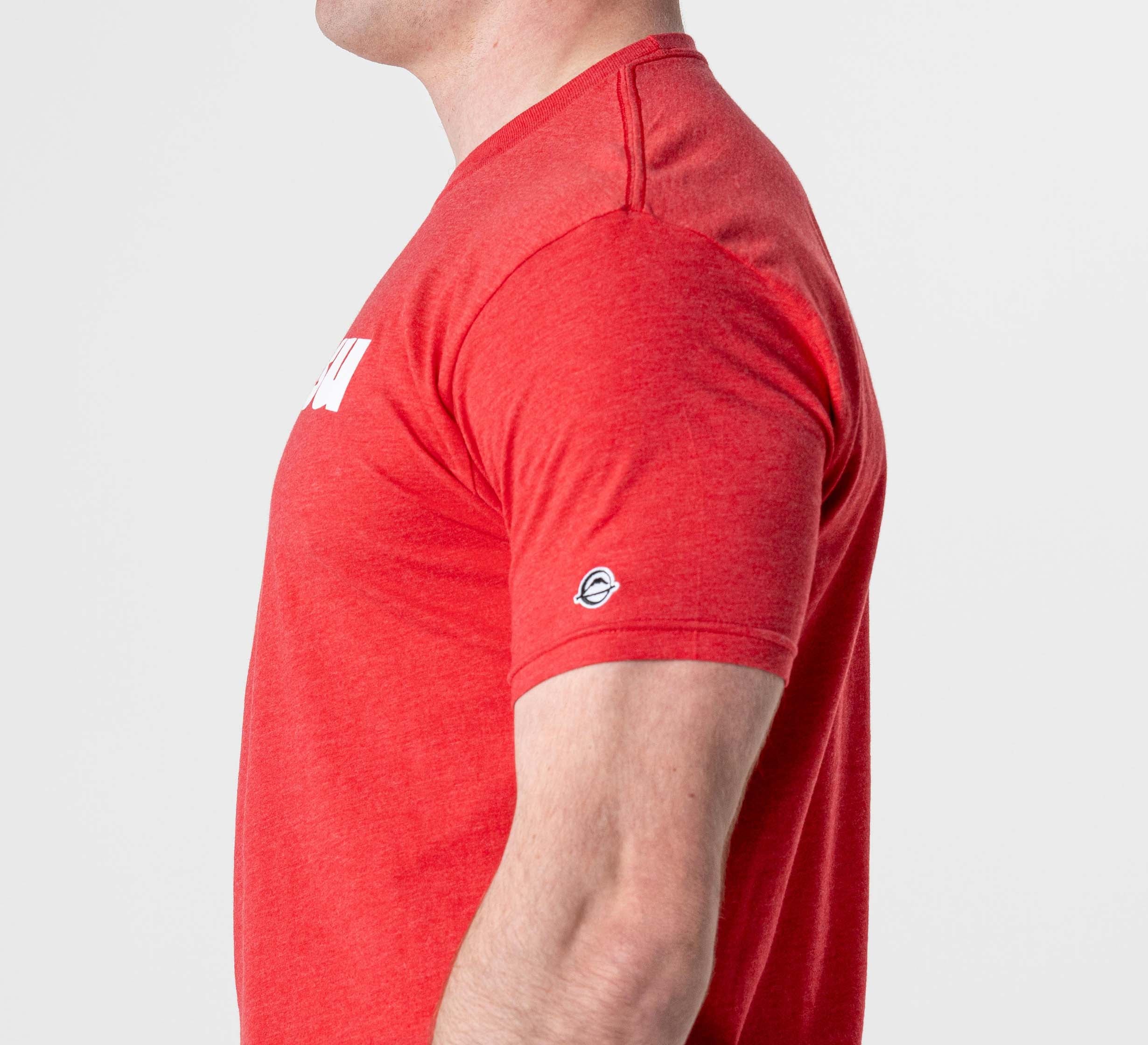 A person is shown from the side wearing the Jiu Jitsu Player T-Shirt Red—a fitted, red short-sleeve tee with a small white logo on the sleeve and partial white text on the chest. The plain, light-colored background highlights the shirt.