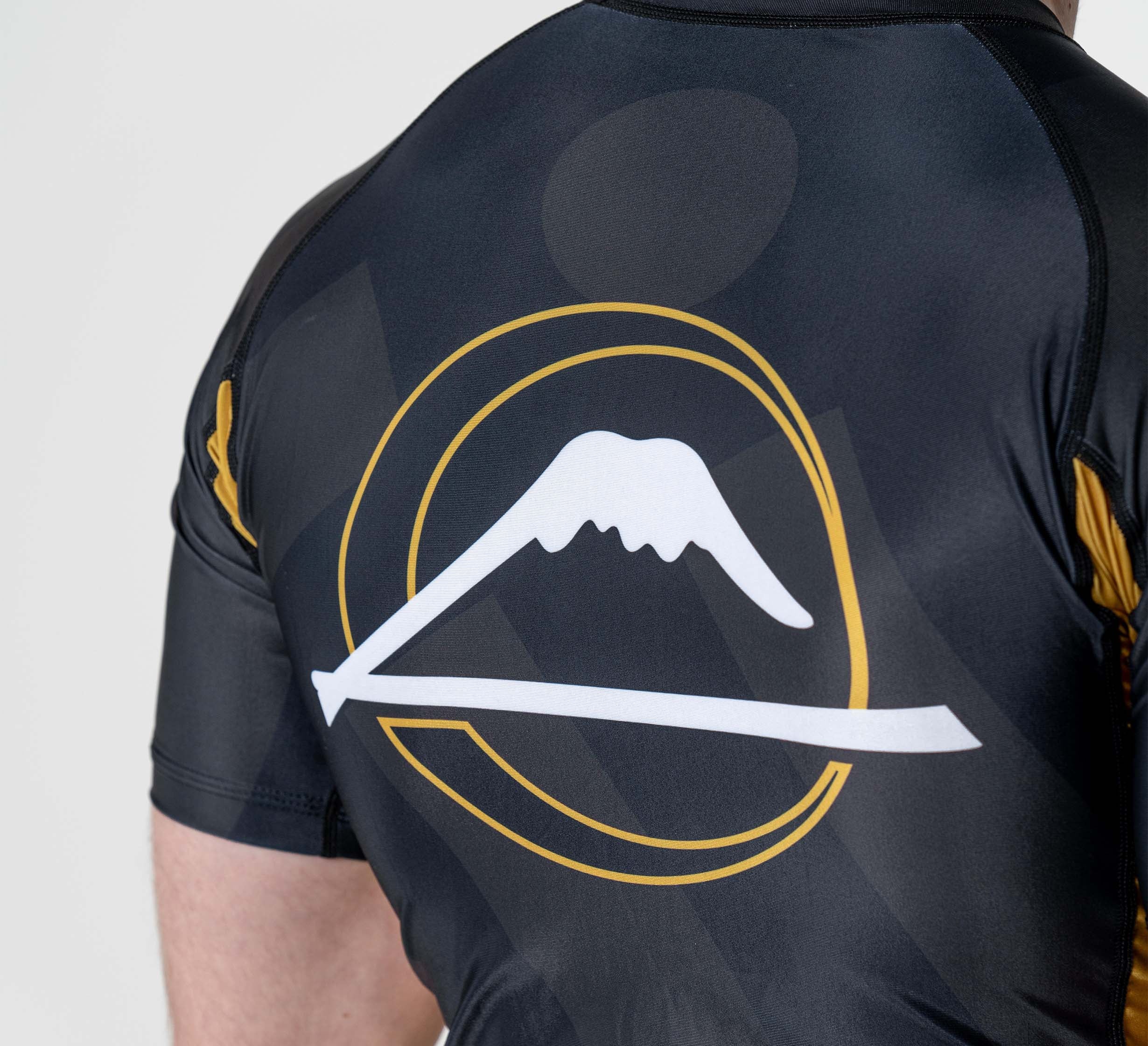 Someone is wearing the Rebel Flex Lite Rashguard Black, which features a stylized logo on the back with a white mountain outline and sword framed by a gold circular line.