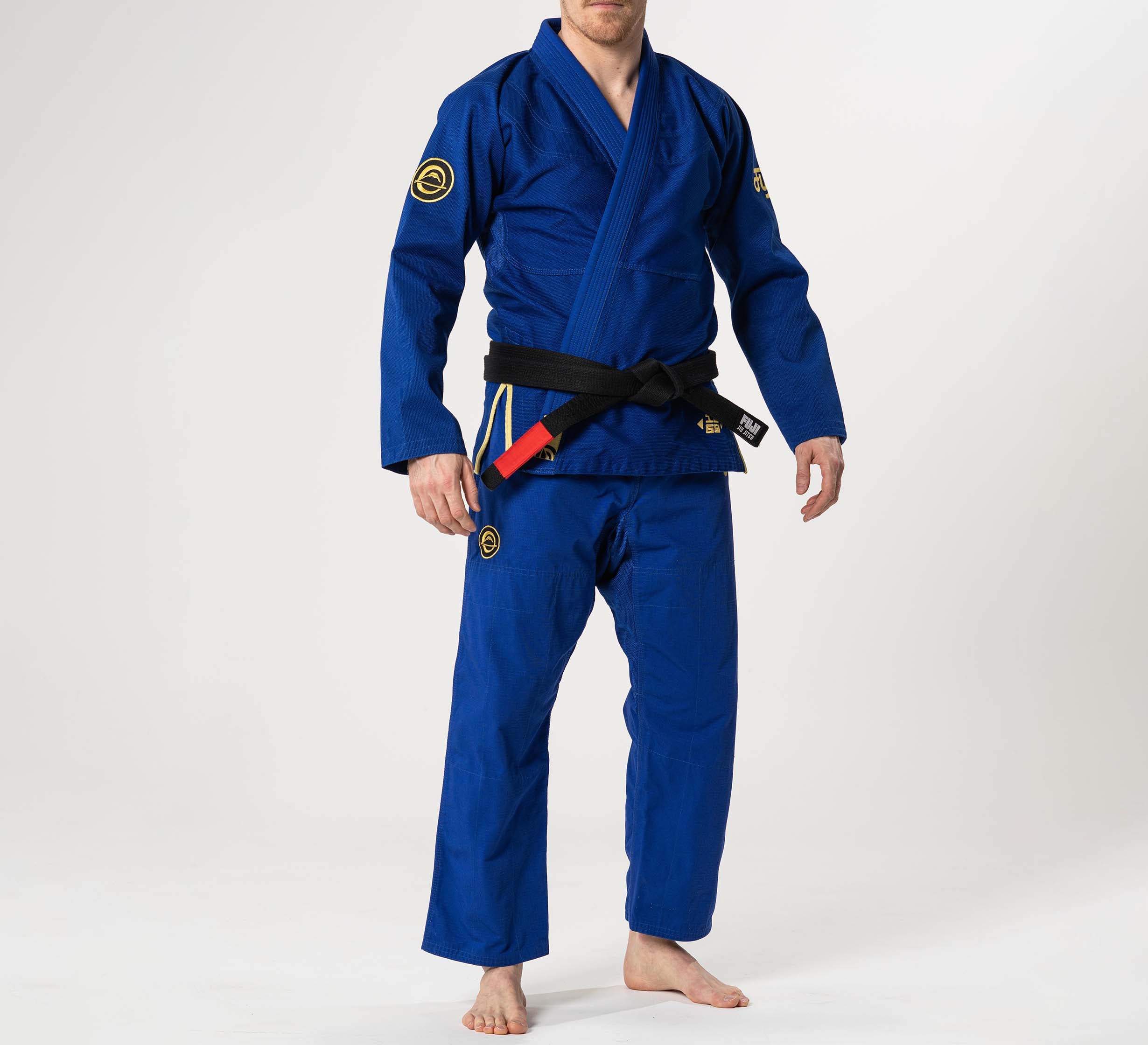 A person wearing the Flow-Tech BJJ Gi Blue/Gold with a black belt, standing barefoot against a plain white background.