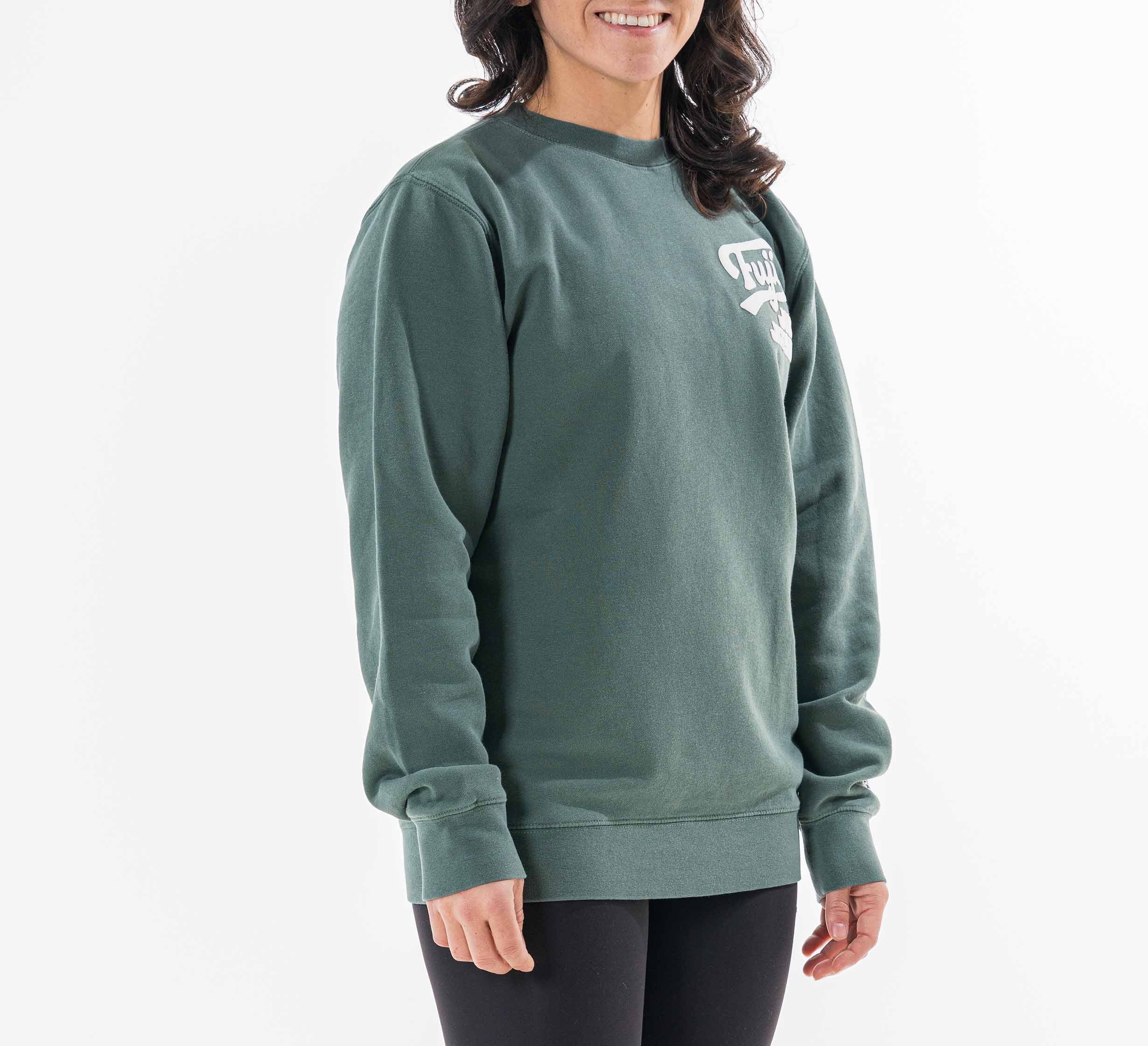 A smiling woman with wavy dark hair wears the Womens Signature Crewneck Green featuring a small white graphic on the left chest, paired with black leggings, against a plain white background.