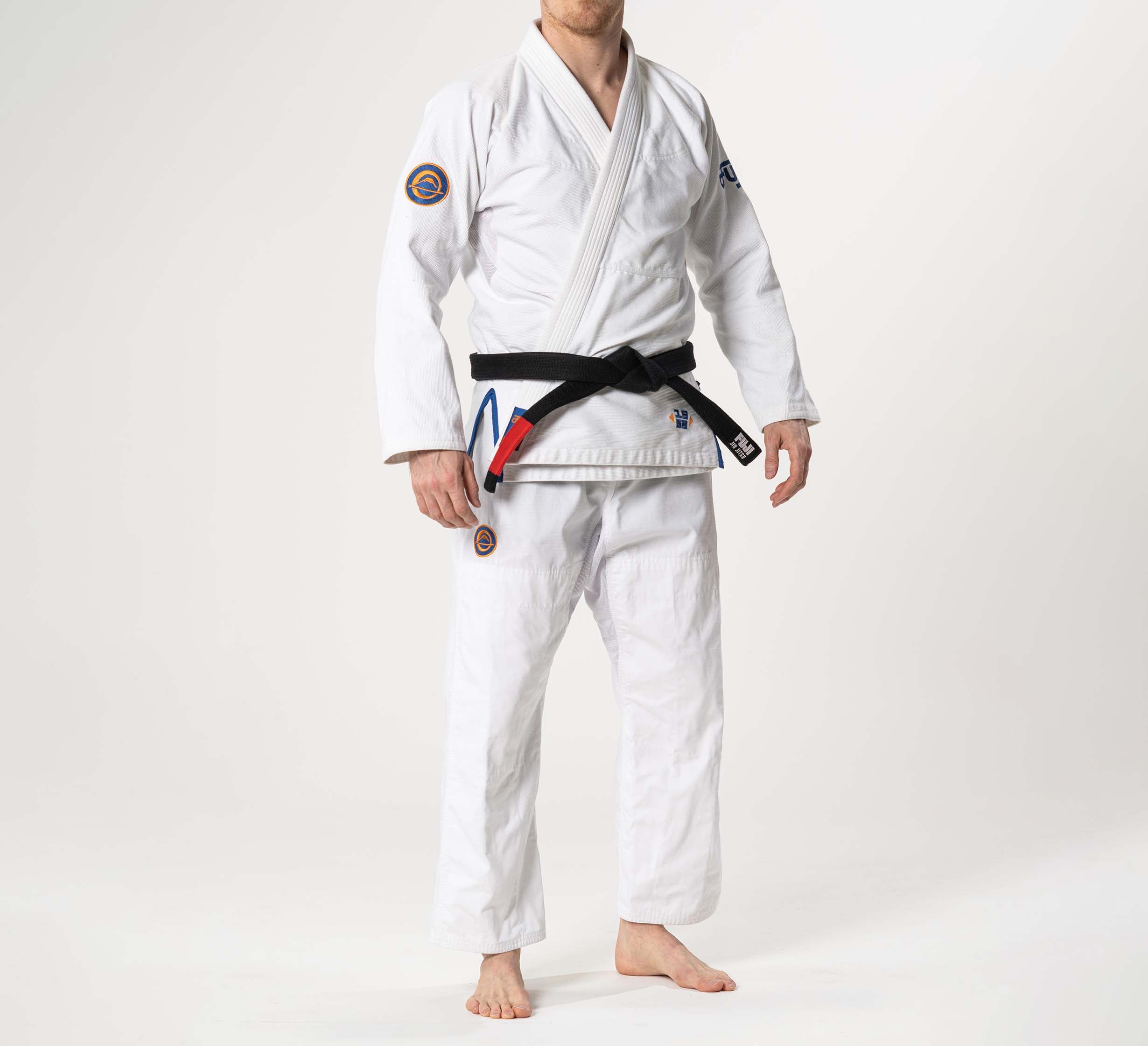 A person stands barefoot in a Flow-Tech BJJ Gi White/Navy/Orange with a black belt and patches, against a plain white background. Their face is not visible.