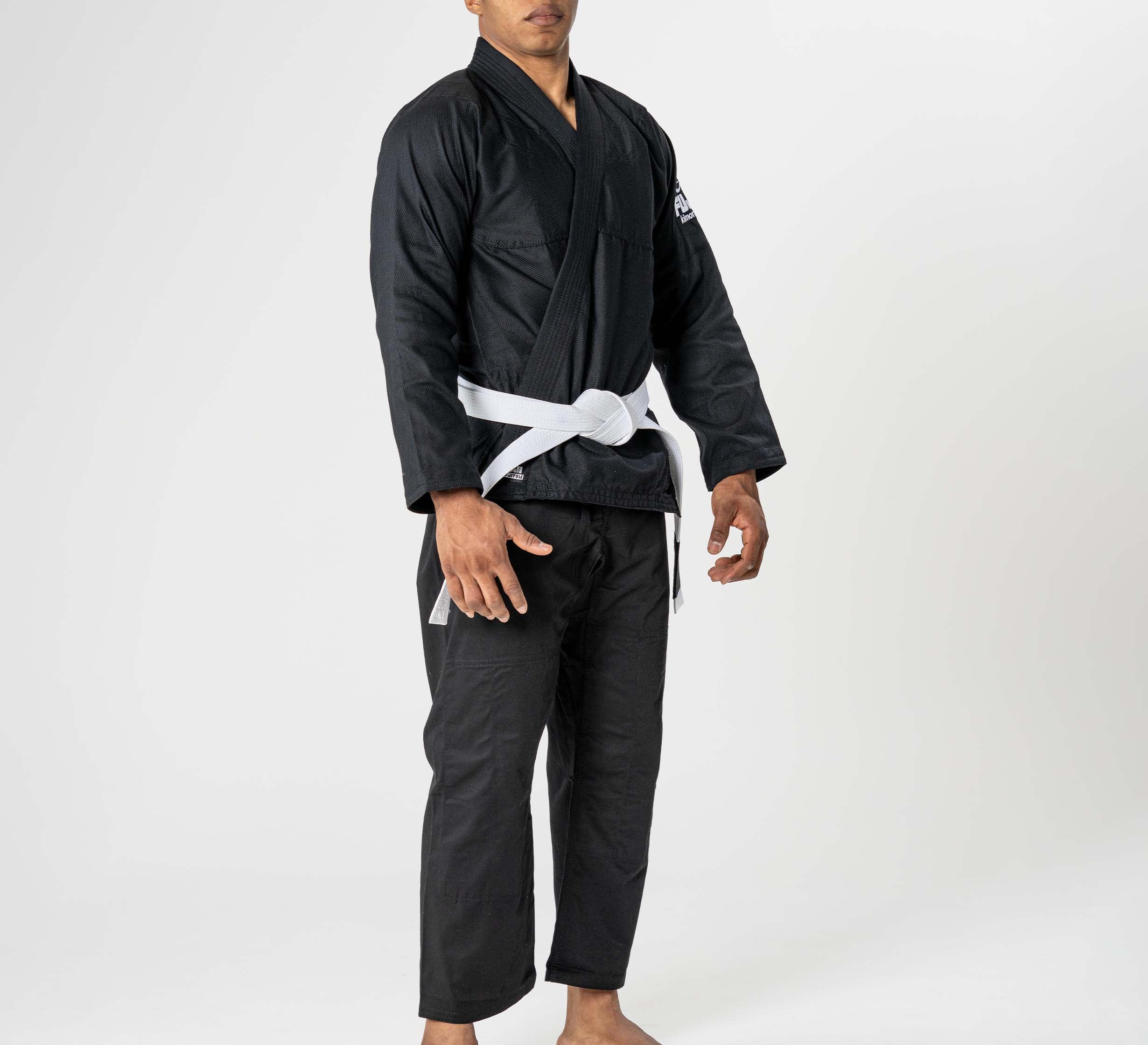 A person stands barefoot against a plain white background, wearing the Starter Adult BJJ Gi Black with a white belt.